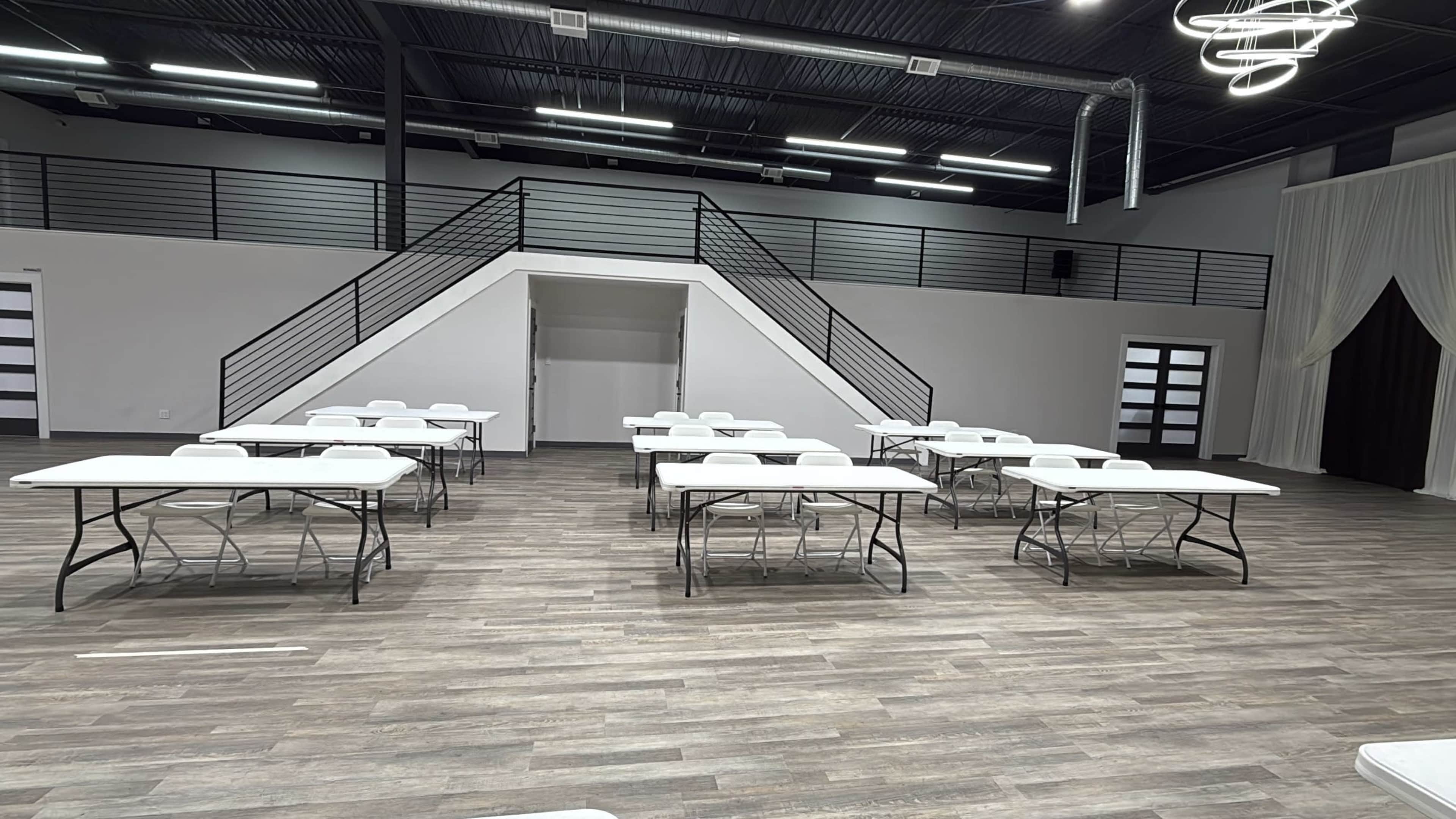The image shows a spacious interior room with several white folding tables arranged in rows and a staircase leading to an upper level.