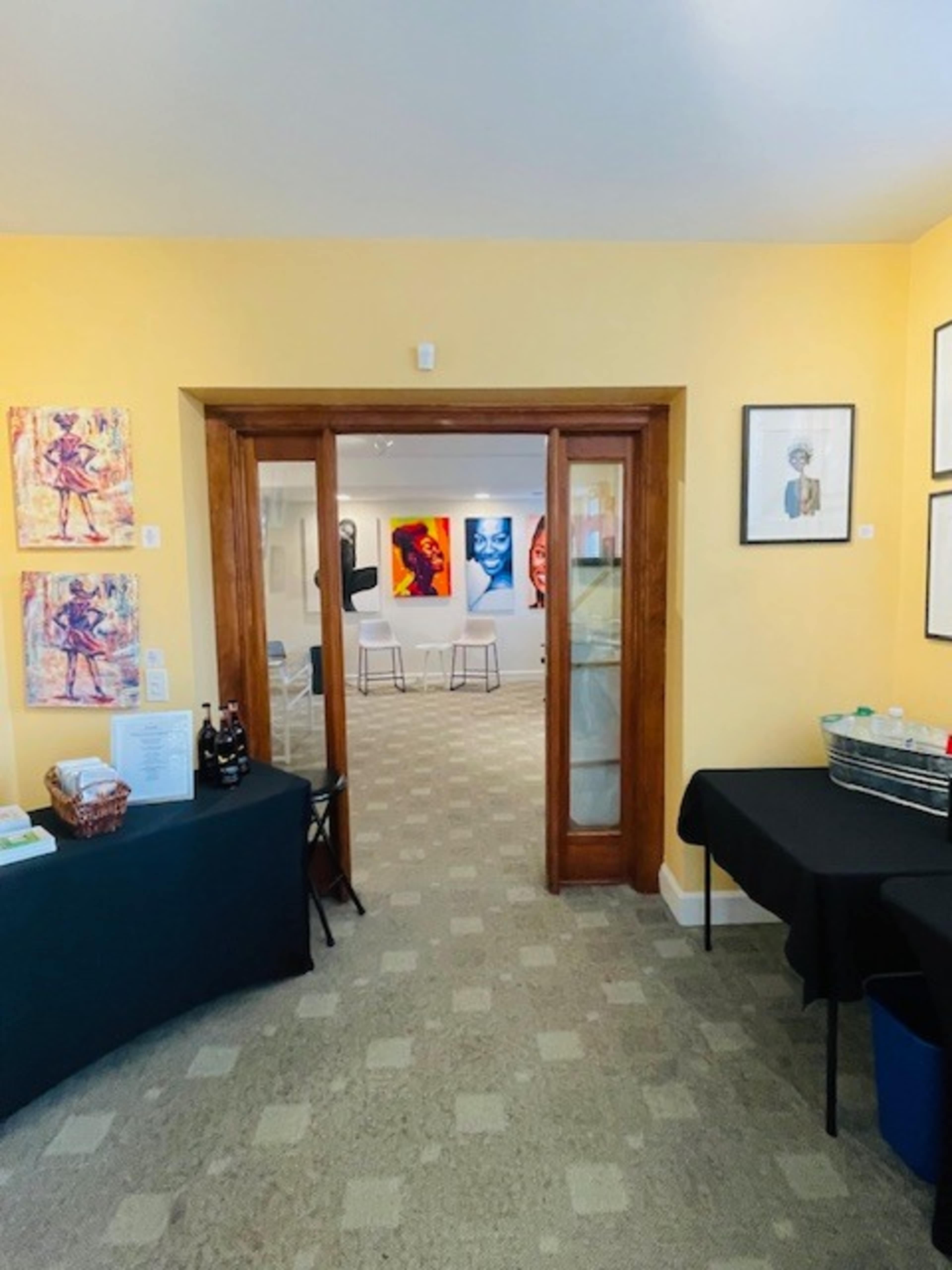 The image shows a doorway leading into an art gallery, with colorful artwork displayed on the walls and a small table on the left side.