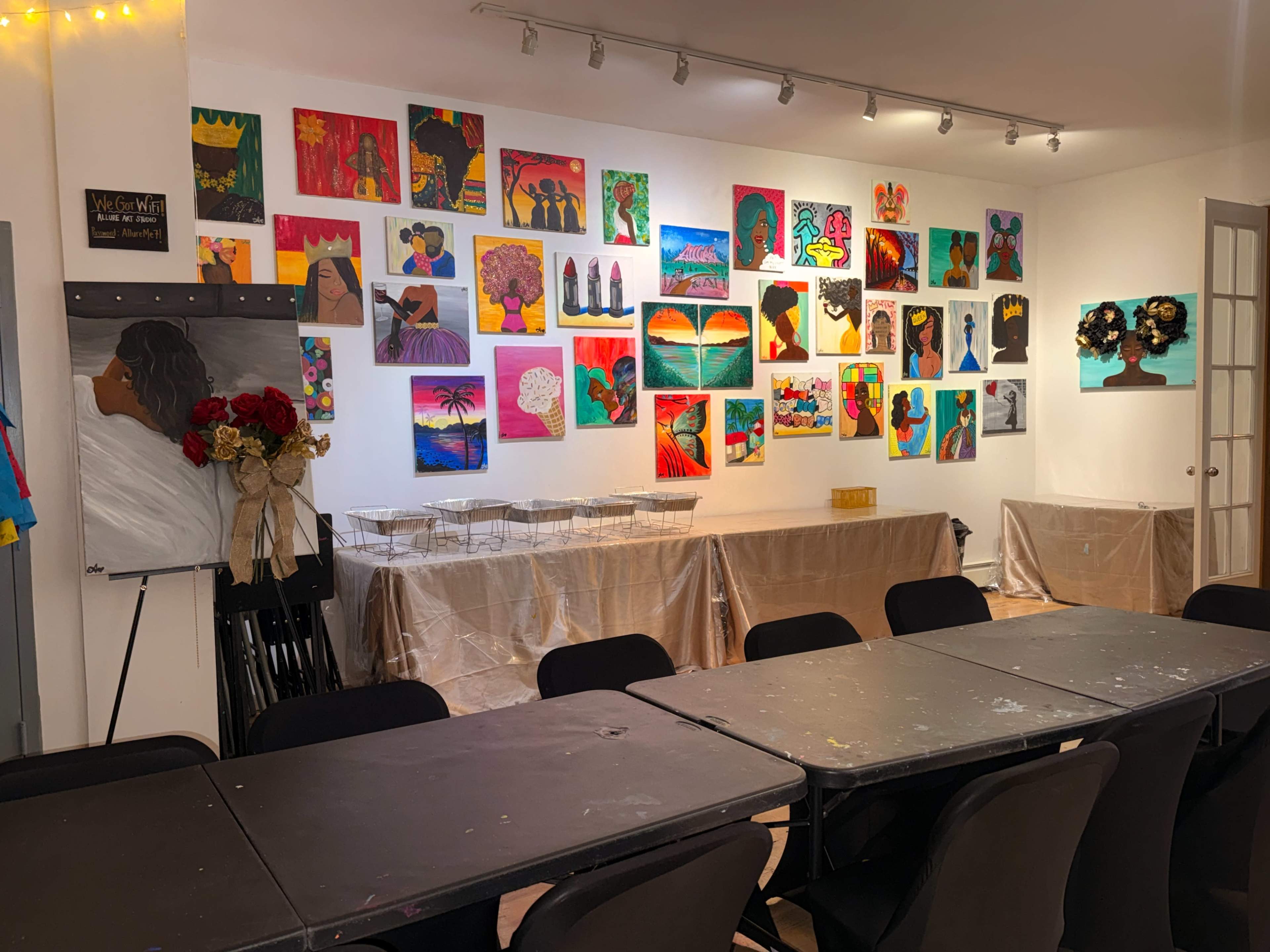The scene features a brightly colored art studio with a wall adorned with various vibrant artworks and a table set up for an event.