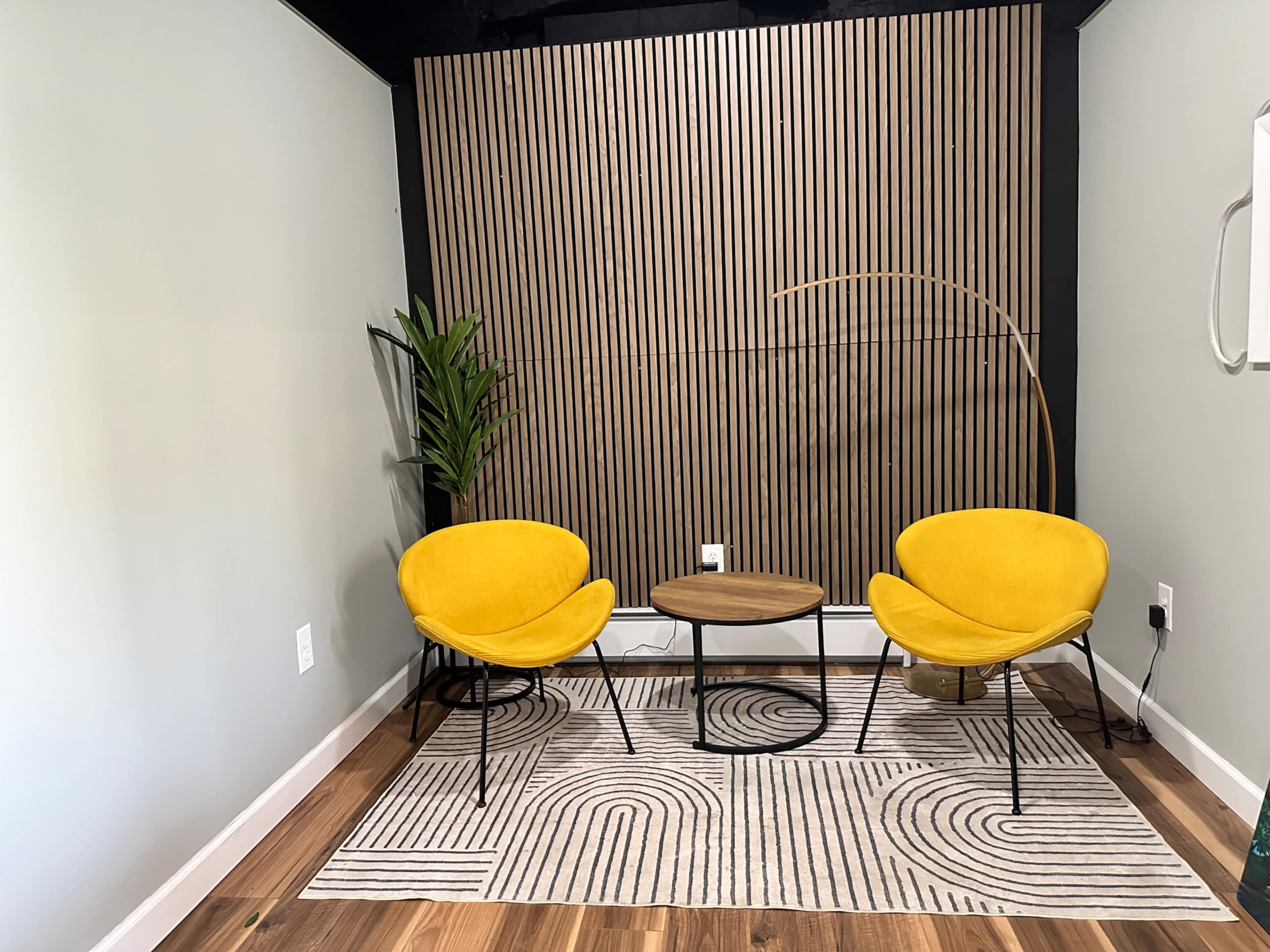 The image shows a cozy seating area with two yellow chairs, a circular wooden table, and a decorative rug against a backdrop of vertical wooden slats.