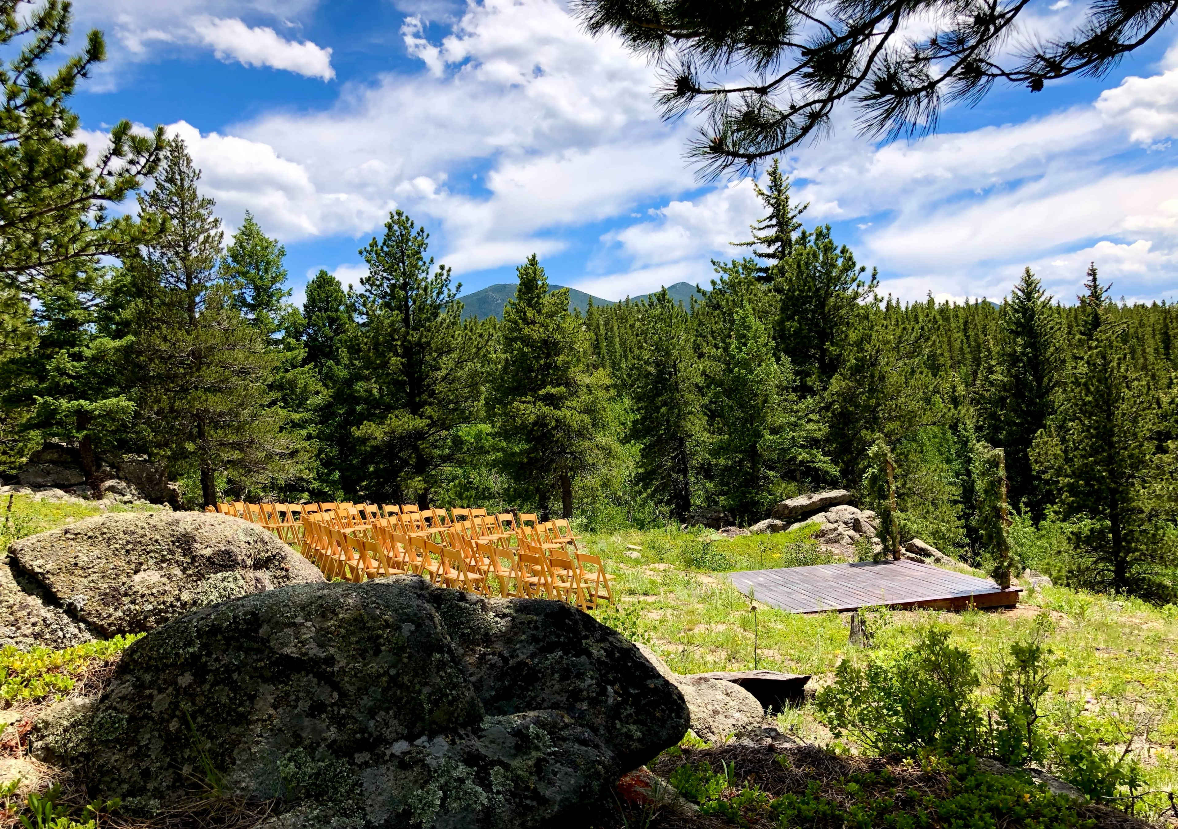 A wooden platform stands in a clearing surrounded by trees, with rows of empty chairs set up for an event.
