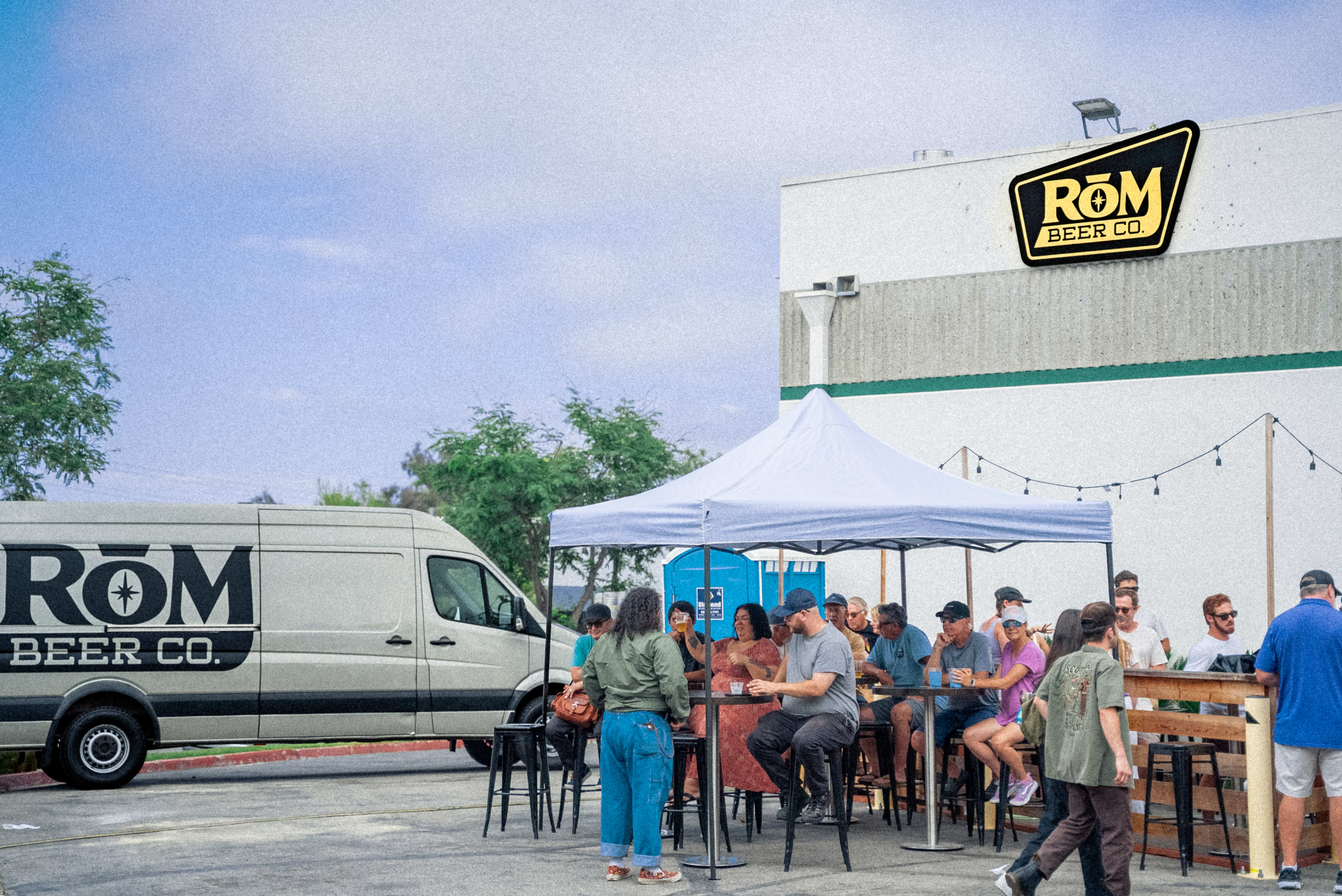 A group of people is seated at outdoor tables near a white building with the sign "RŌM BEER CO," while a delivery van is parked nearby.
