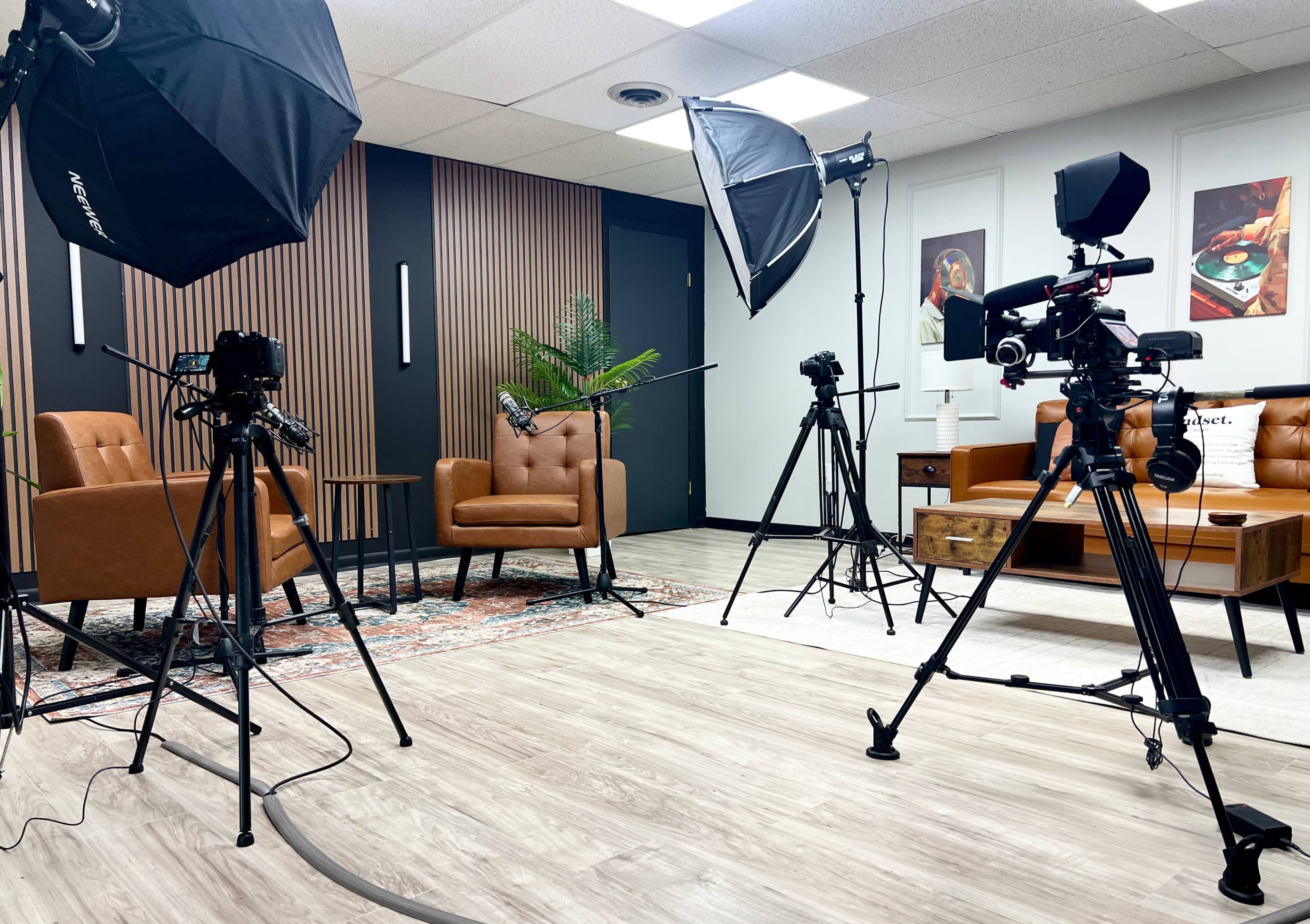 The image shows a well-lit interview setup with two brown chairs, cameras on tripods, softbox lights, and a decorative plant in a modern office space.