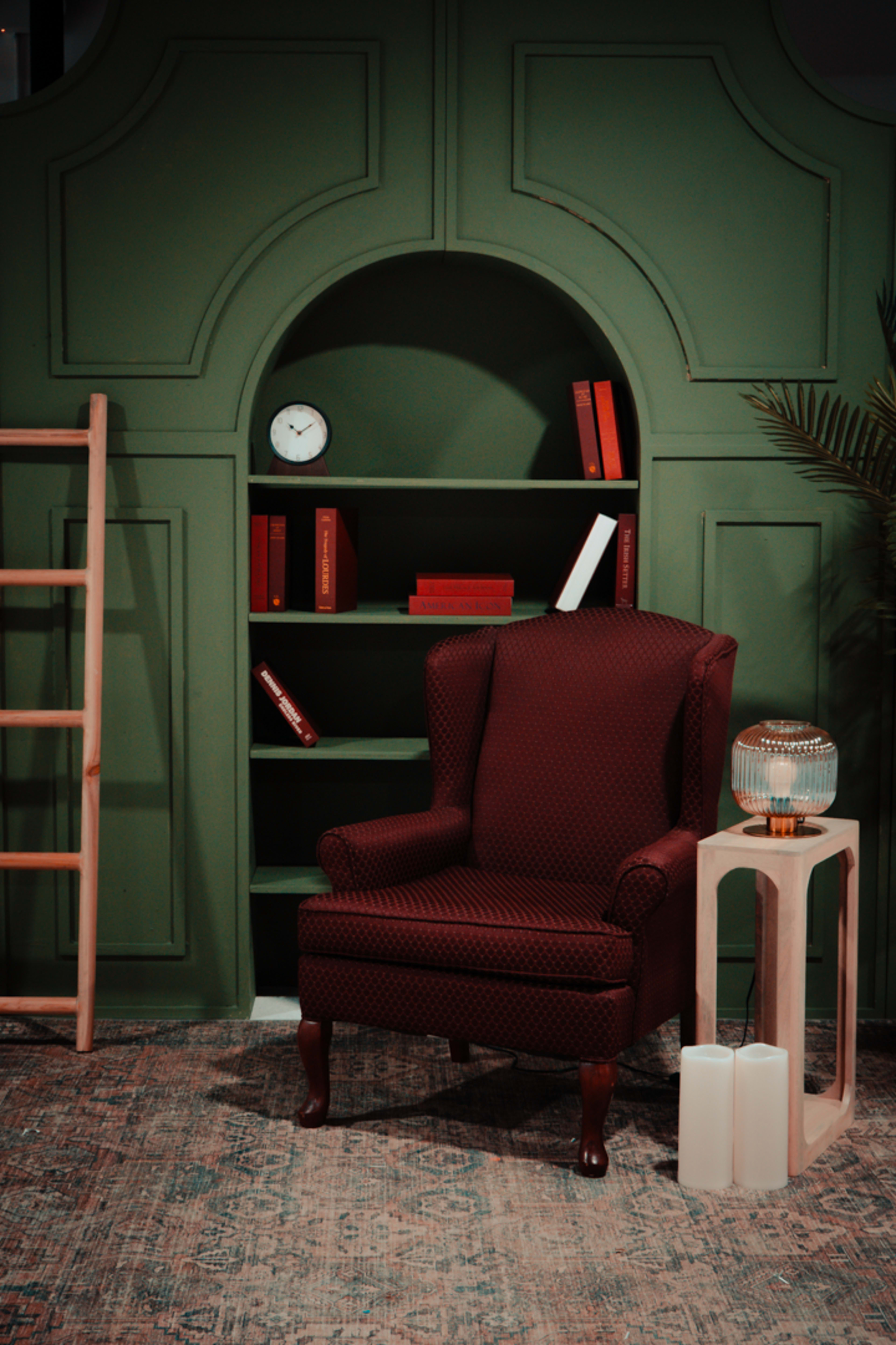 A burgundy upholstered armchair sits next to a side table with a glass lamp, in front of a green wall featuring shelves filled with books and a clock.