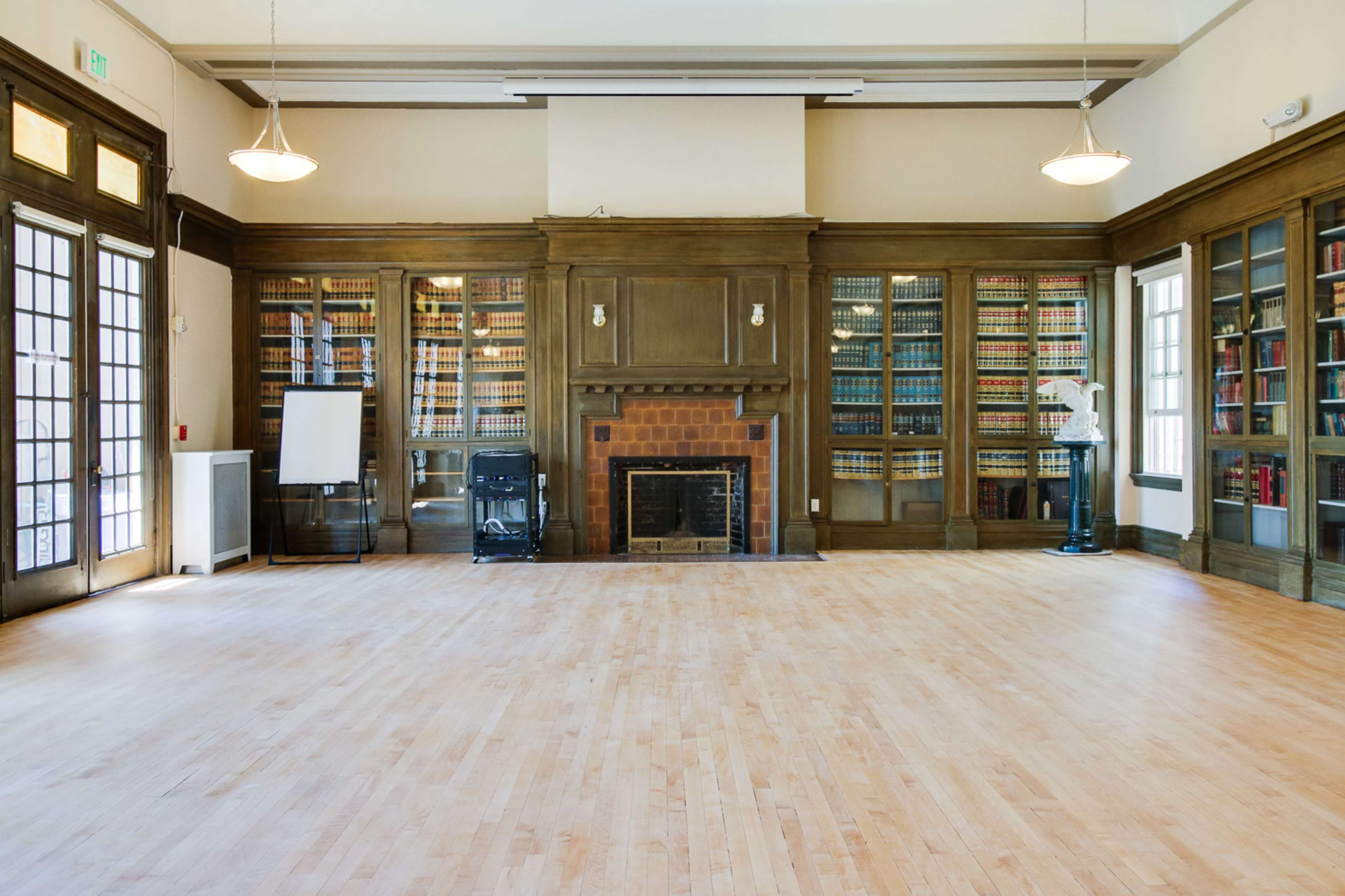 A spacious room with wooden flooring, flanked by bookshelves filled with books on either side, and featuring a central fireplace and a blank presentation board.