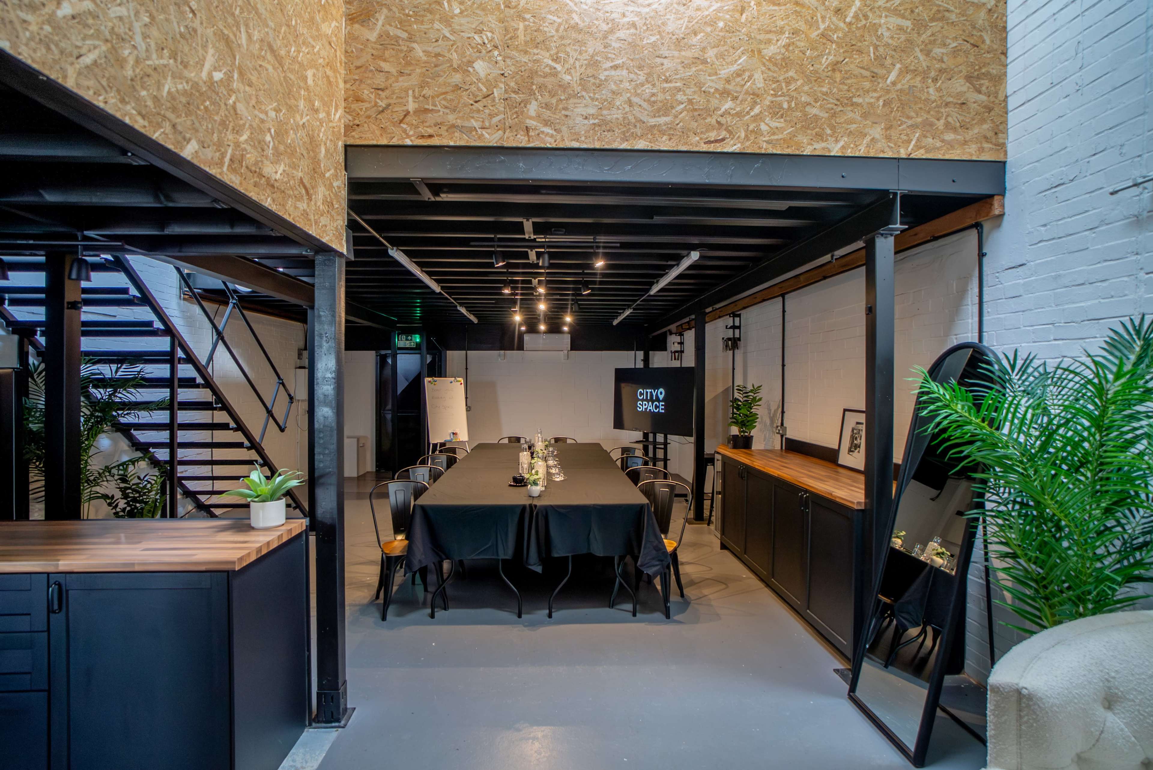 A modern meeting space features a long black table set with plants and decor, surrounded by chairs, with exposed beams and white brick walls.