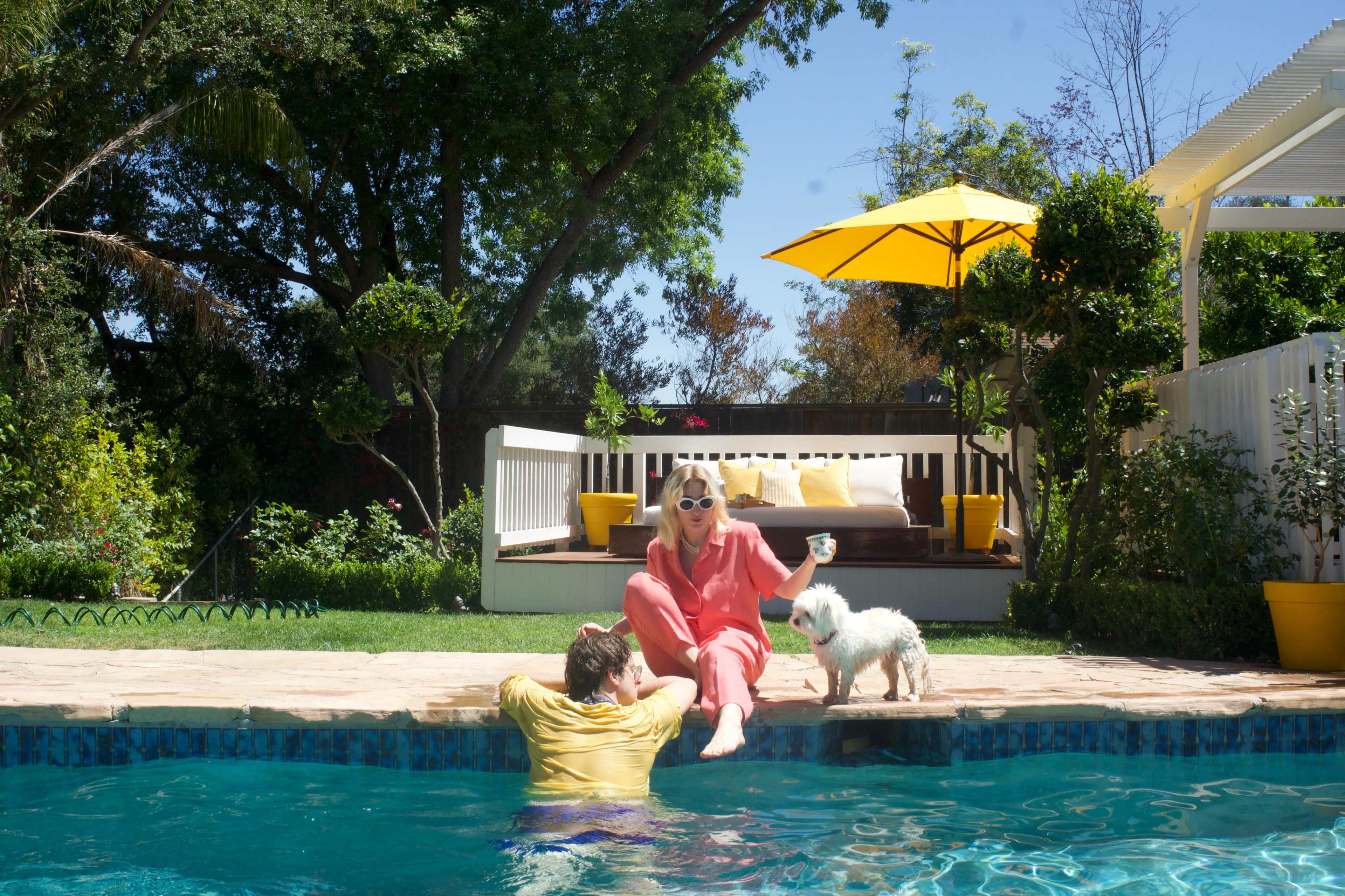 A woman in a pink outfit sits at the edge of a pool while a man partially submerged in the water looks up, and a small white dog stands nearby under a yellow umbrella.