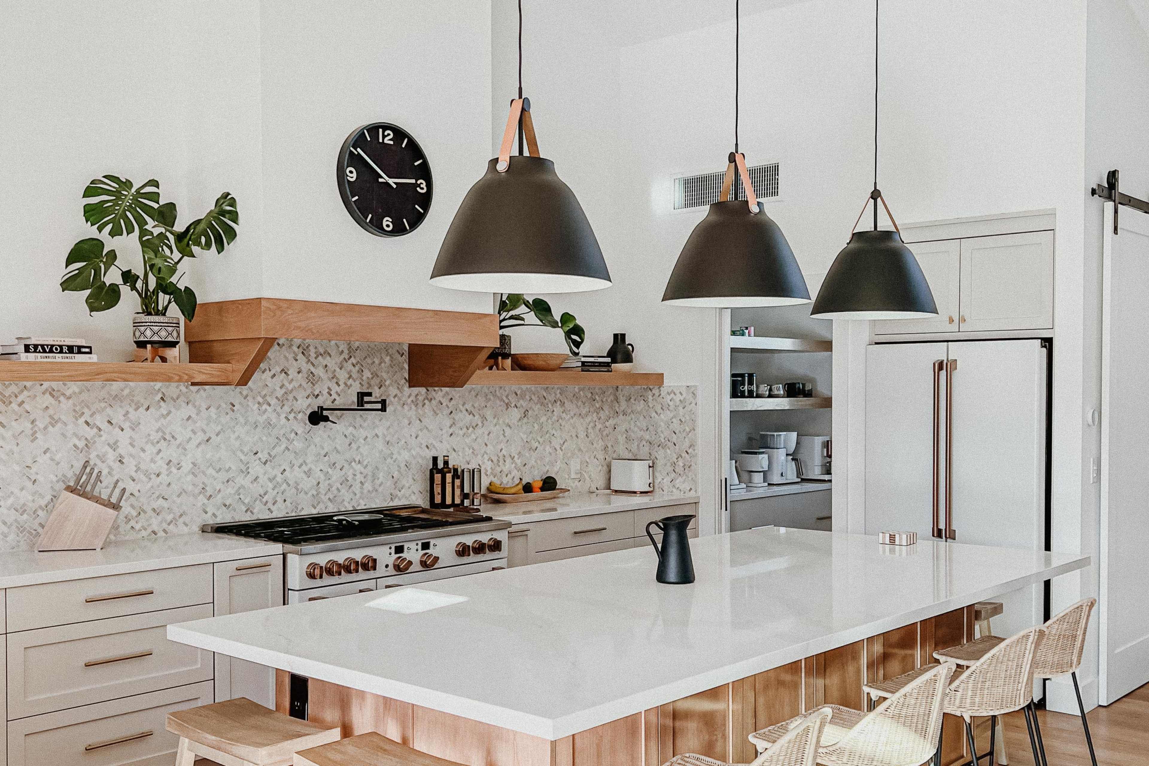 A modern kitchen features a large island with bar stools, a sleek stove, overhead pendant lights, and greenery on the countertop.