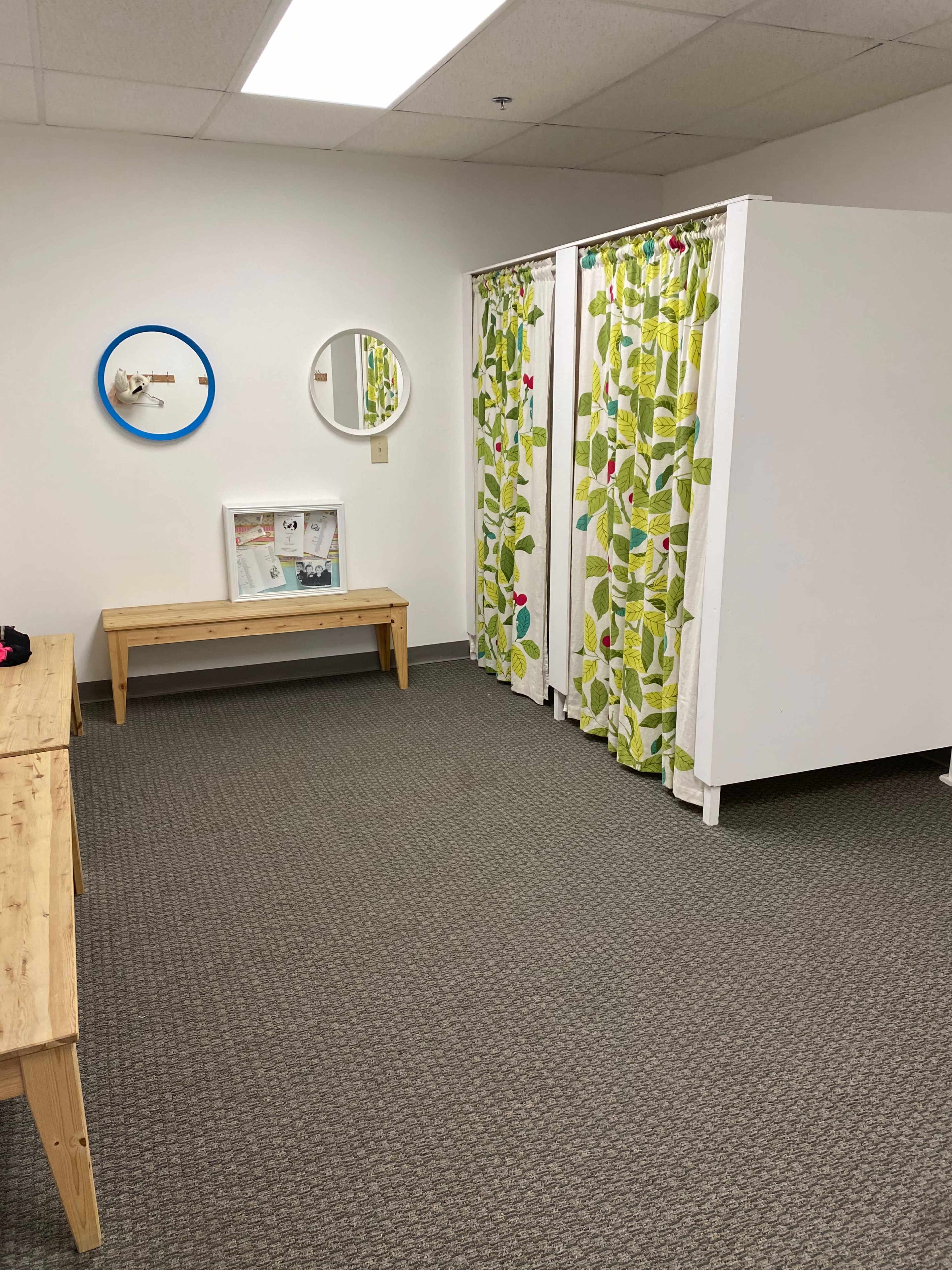 The image shows a minimalistic fitting room area with two white curtain-enclosed stalls, a wooden bench, and two round mirrors on the wall.