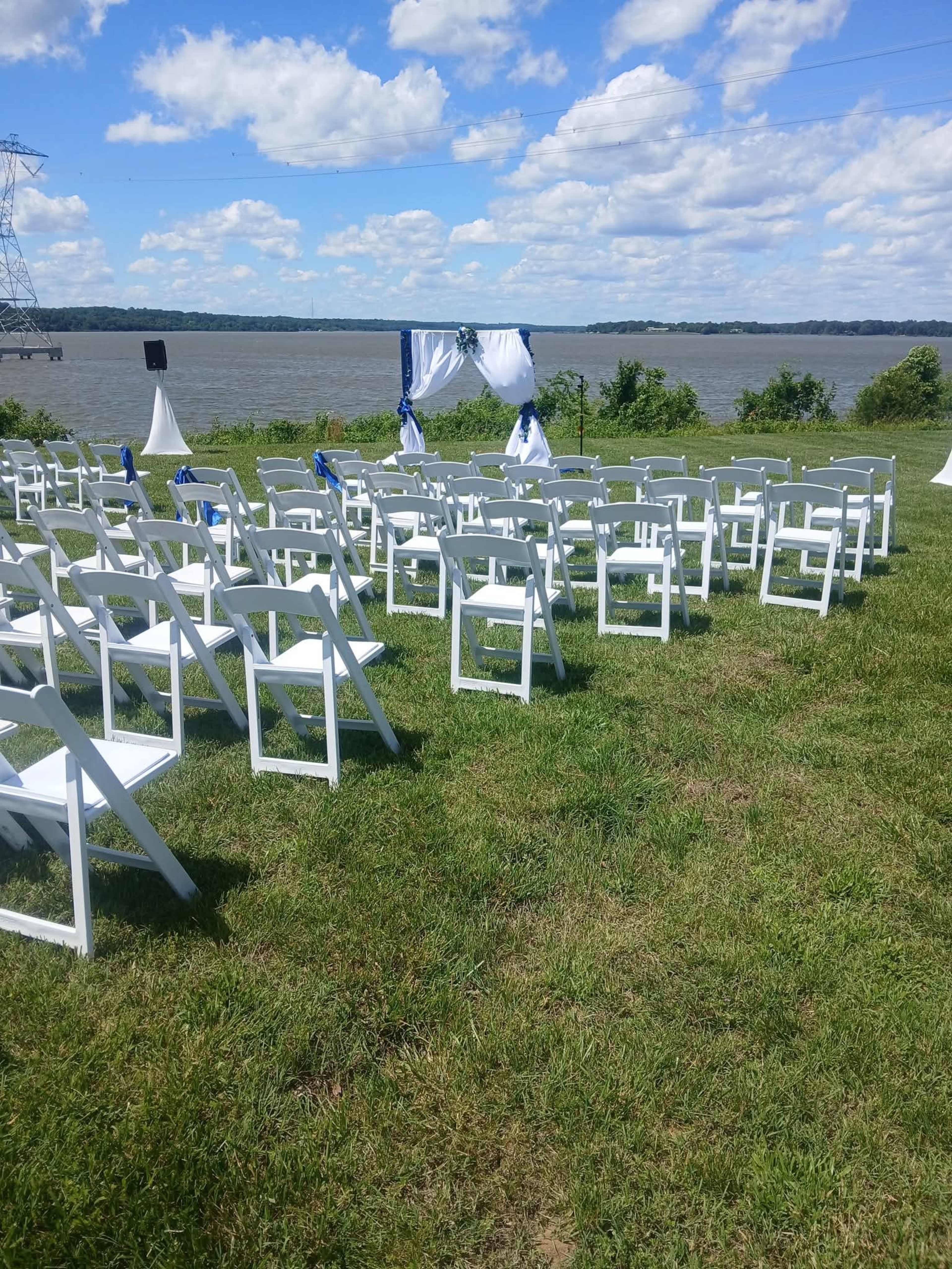 A row of white chairs is arranged on a grassy area facing a decorative arch near a river, under a partly cloudy sky.