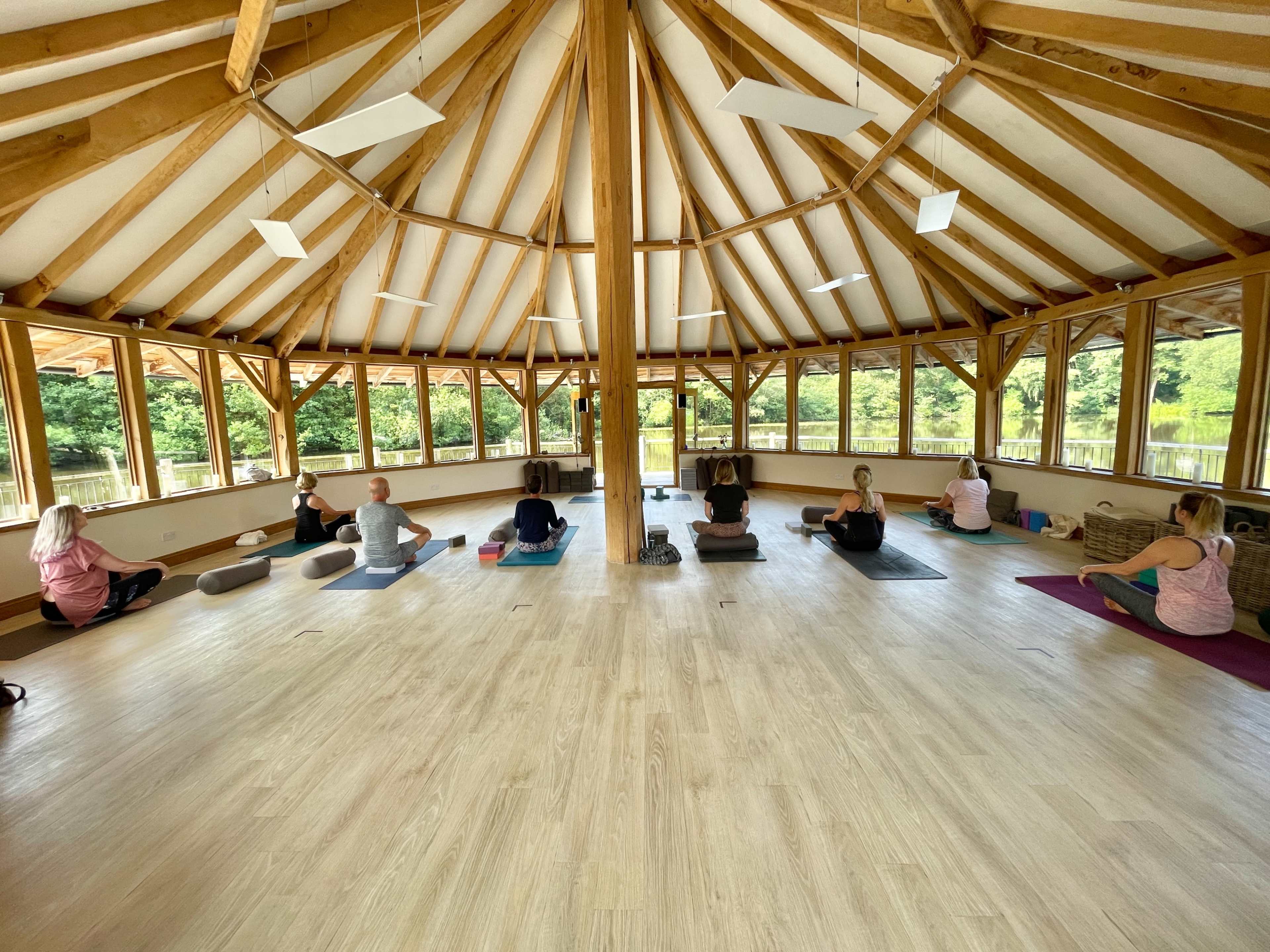 A group of people are practicing yoga in a spacious, wooden studio with large windows overlooking greenery.