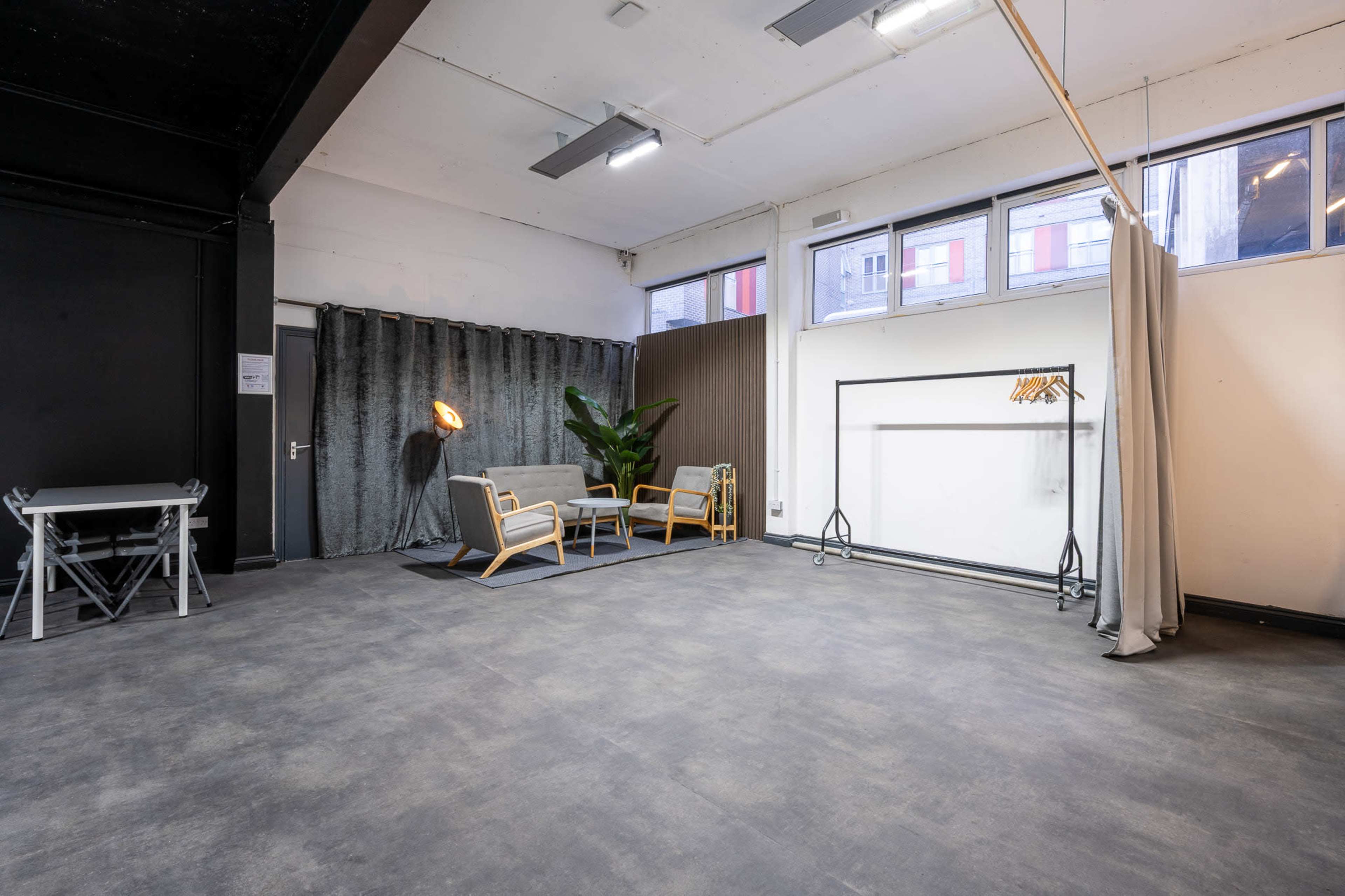 The image shows an empty studio space featuring a seating area with two chairs, a sofa, and a large curtain, along with a table and a standing lamp.