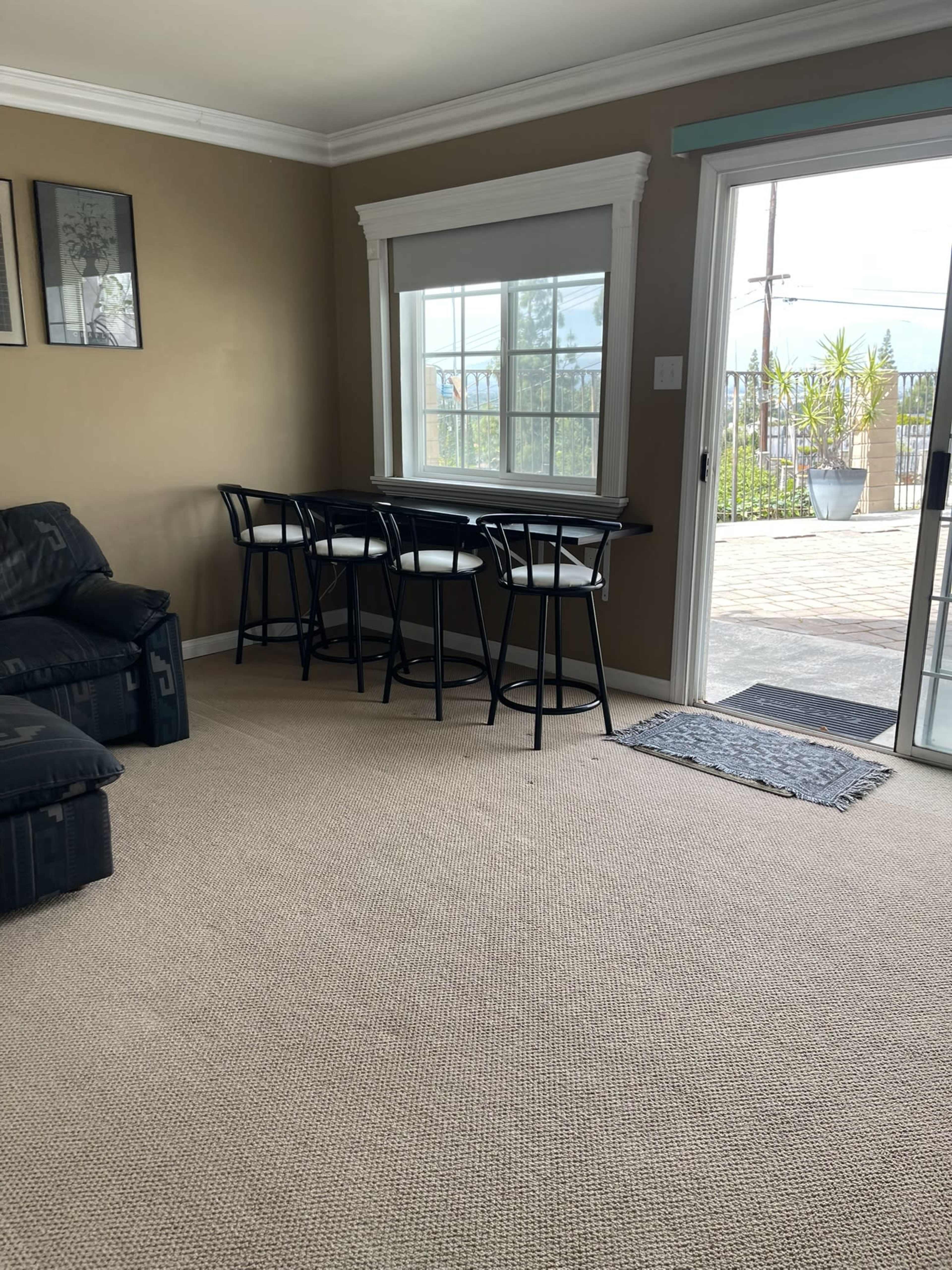 The image shows a carpeted living area with a black couch, a small table with four black chairs, and a sliding glass door leading to an outdoor space.