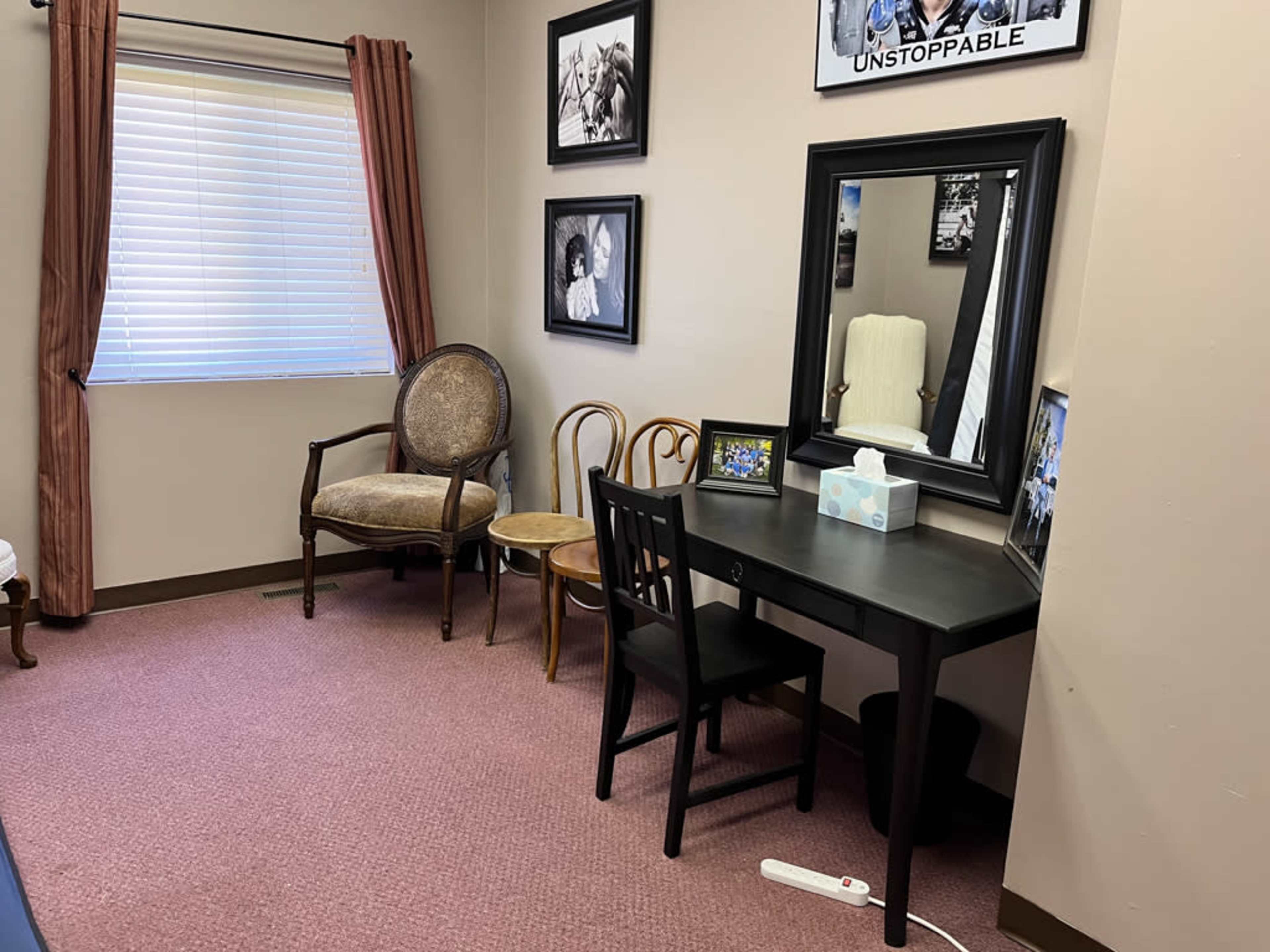 A small room with a black desk and chair, a round armchair, two wooden chairs, a mirror, and several framed photos on the walls near a window with closed blinds.