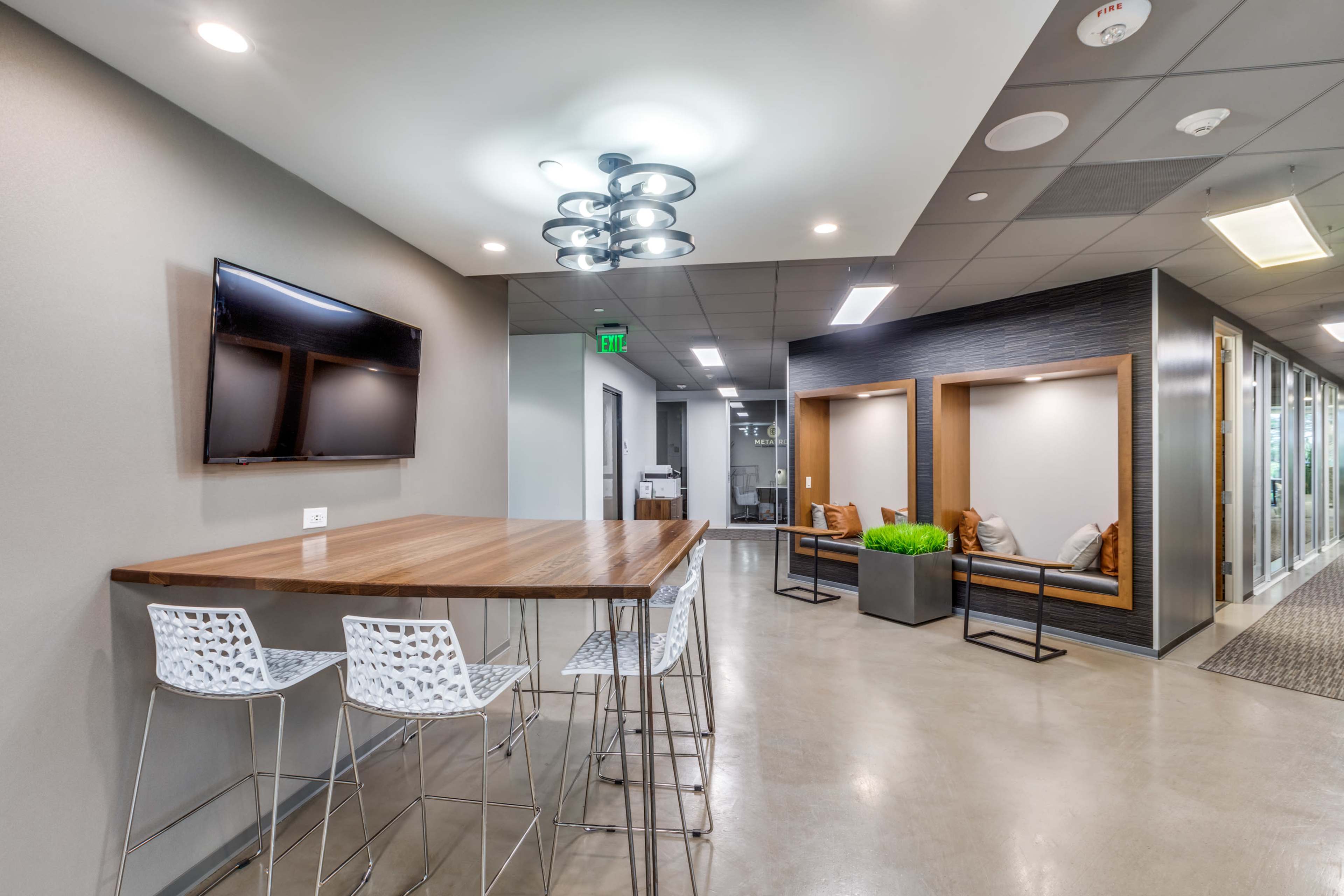 The image shows a modern office space featuring a sleek wooden table with bar stools, a wall-mounted TV, and a waiting area with seating along a corridor.