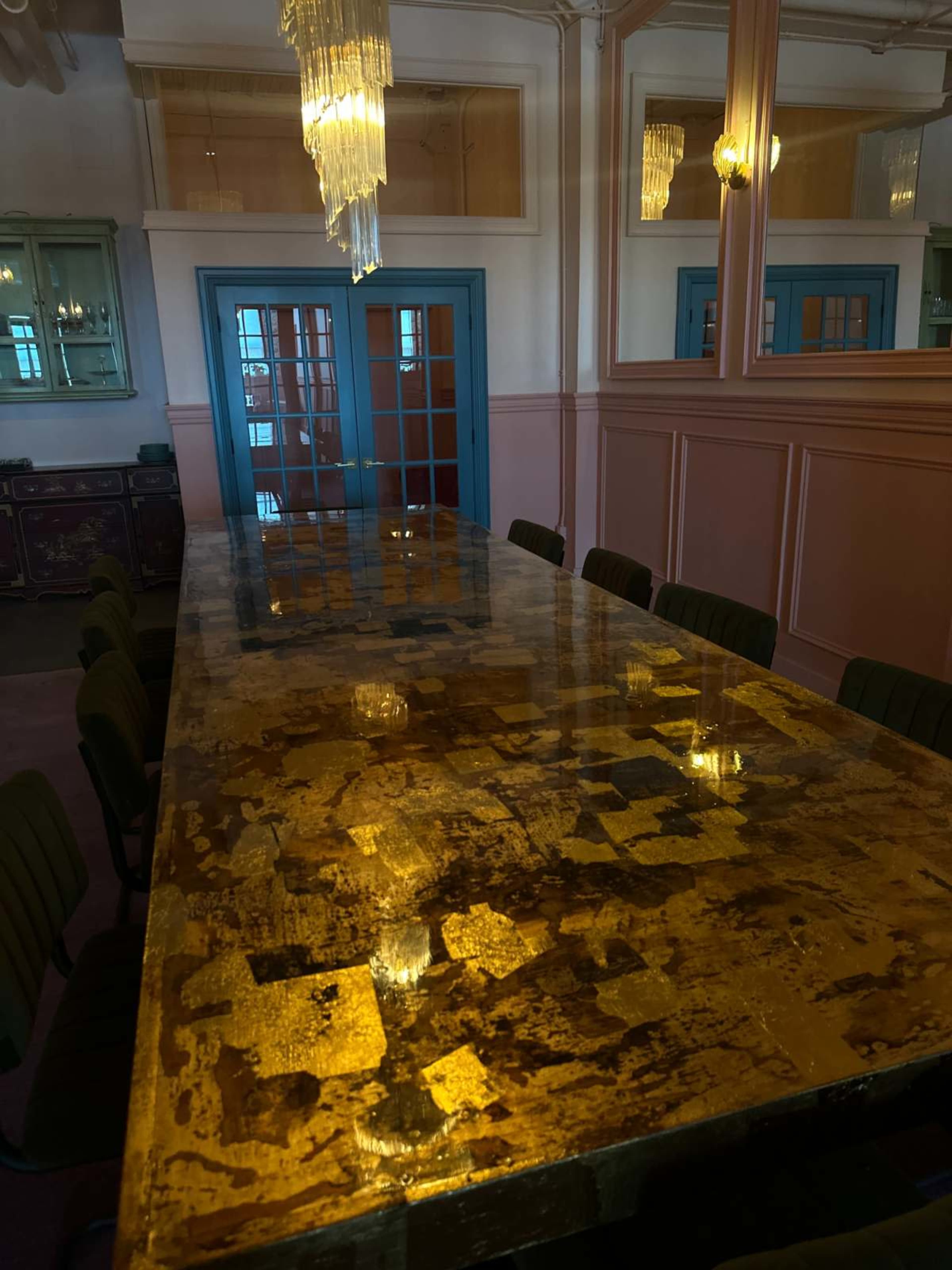 A long table with a glossy, gold-patterned surface is centered in a well-lit room adorned with chandeliers and large mirrors.