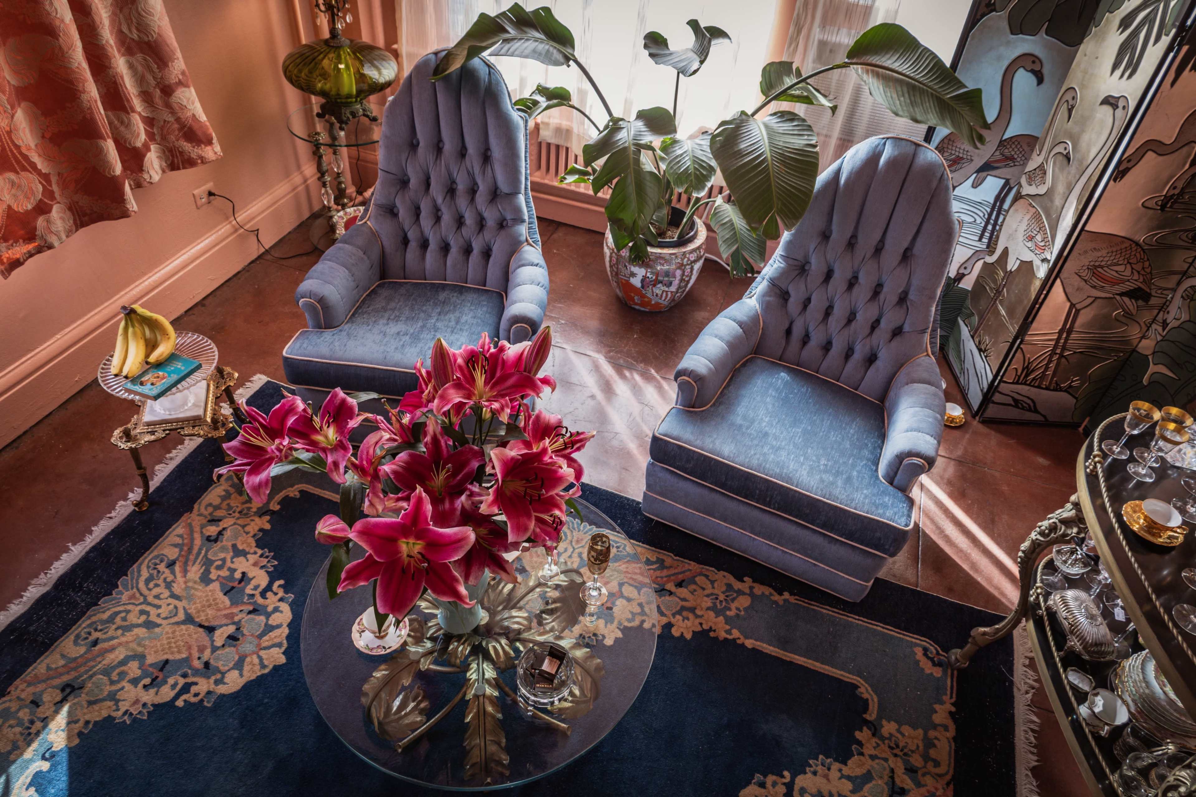 The image depicts two blue upholstered armchairs flanking a round glass table adorned with a floral arrangement of pink lilies and various decorative items in a stylized interior.