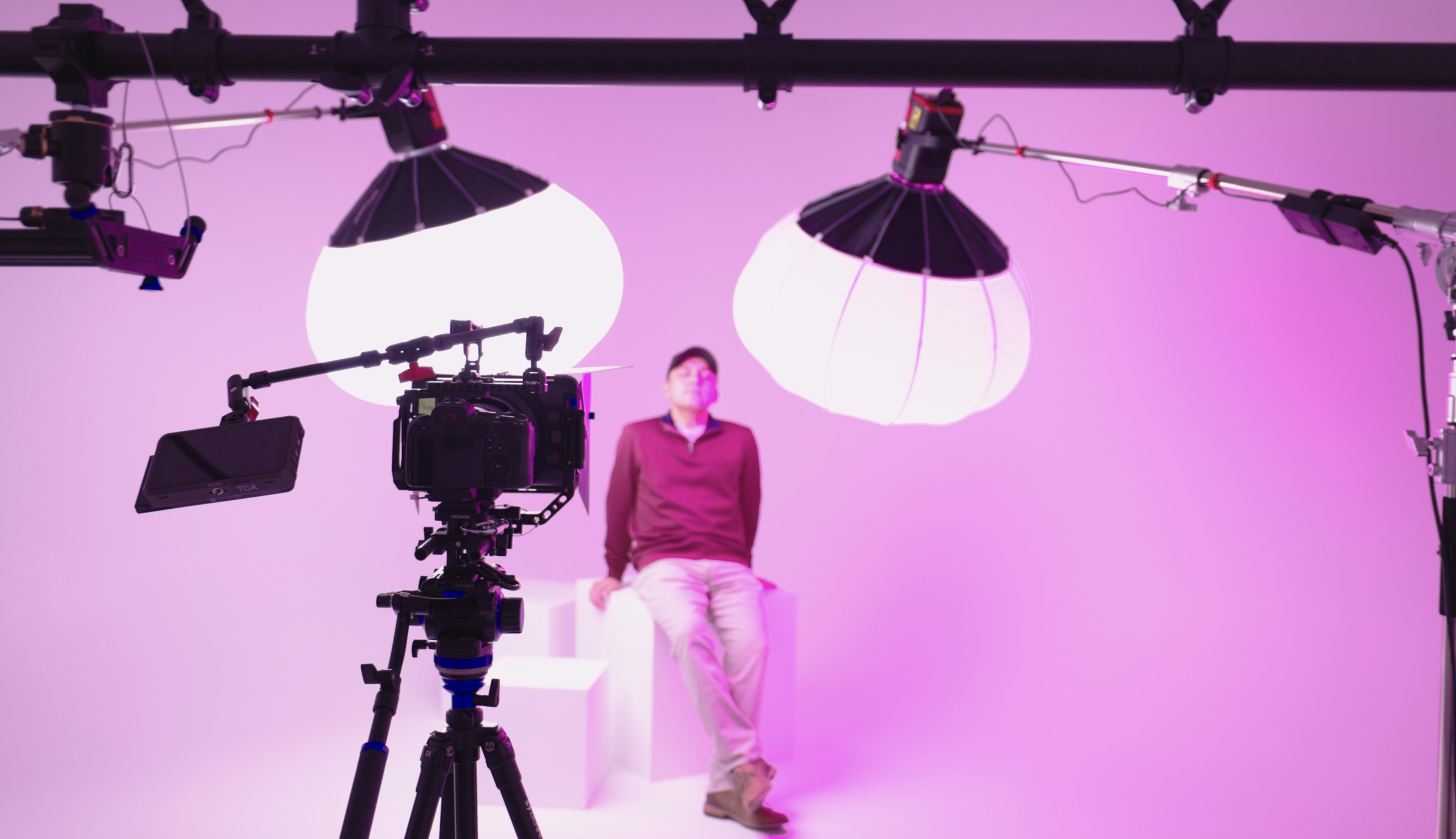 A camera on a tripod is positioned in front of a person sitting on a pedestal under two large, pink-lit softboxes.