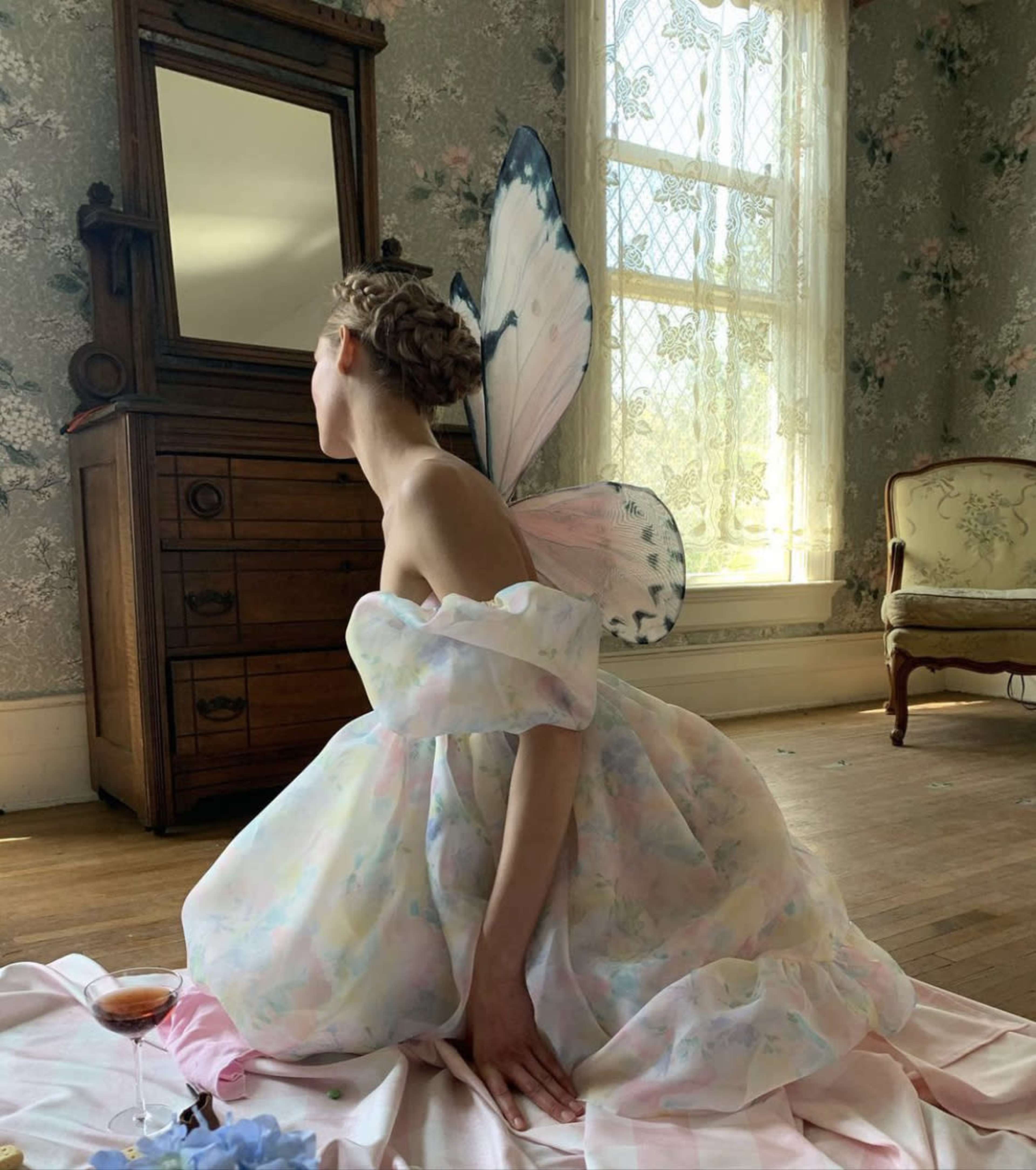 A woman in a pastel-colored dress with large butterfly wings sits on a blanket in a vintage-inspired room, gazing at her reflection in a mirror.