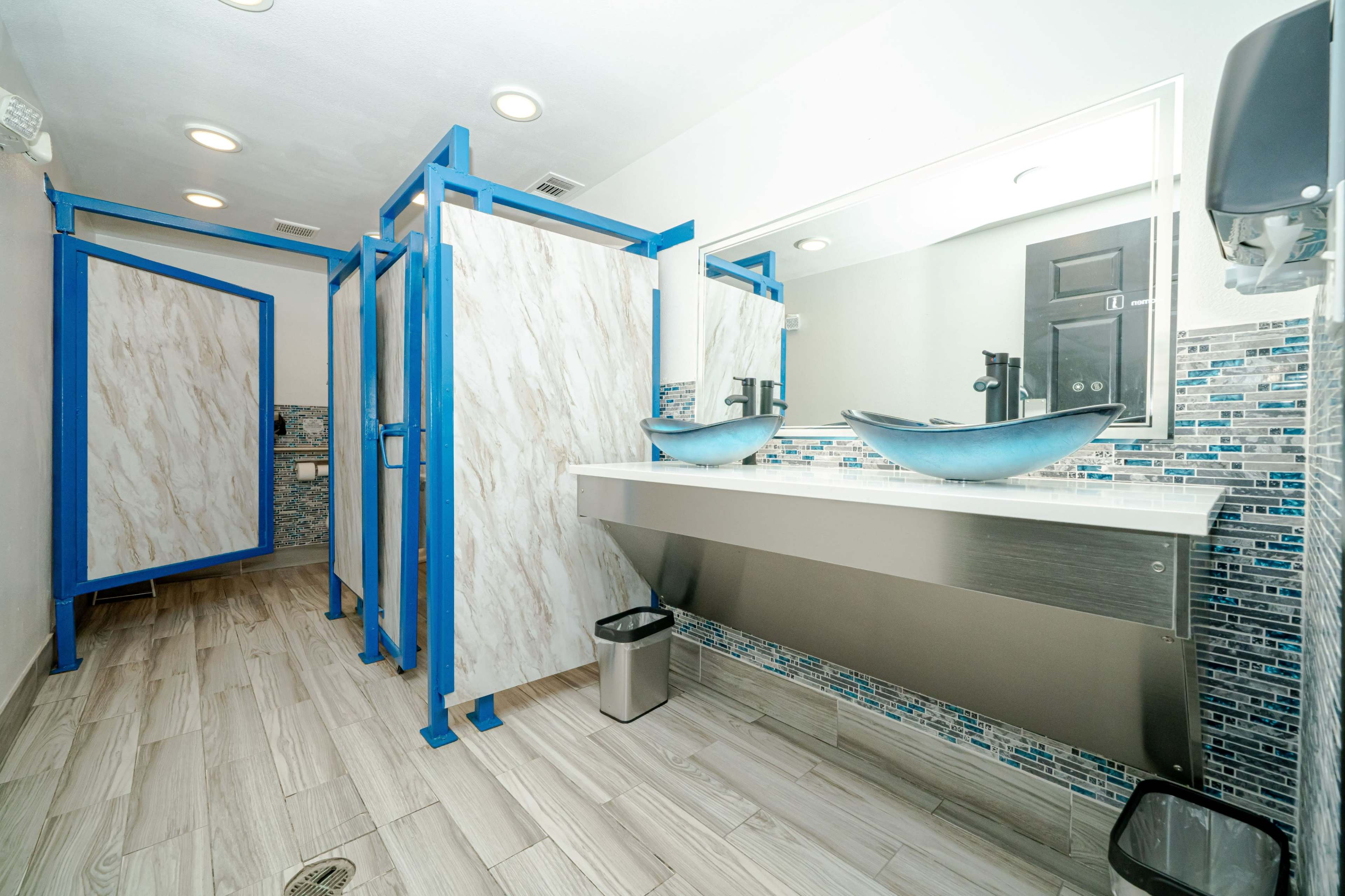 The image shows a modern restroom with three stalls, a long counter with two vessel sinks, and tiled floors with a blue and white color scheme.