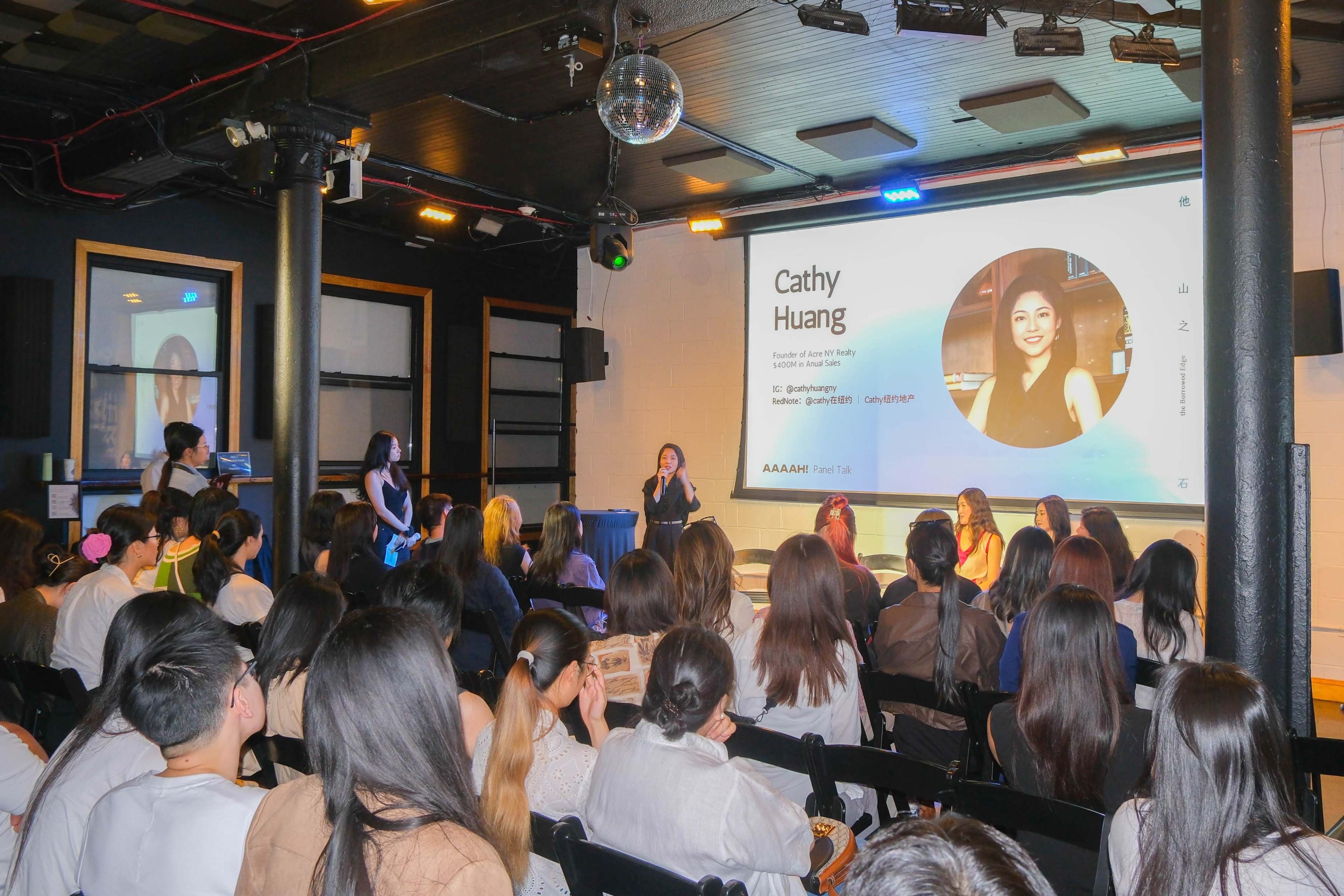 A speaker stands on stage in front of an audience during a panel discussion or seminar event.
