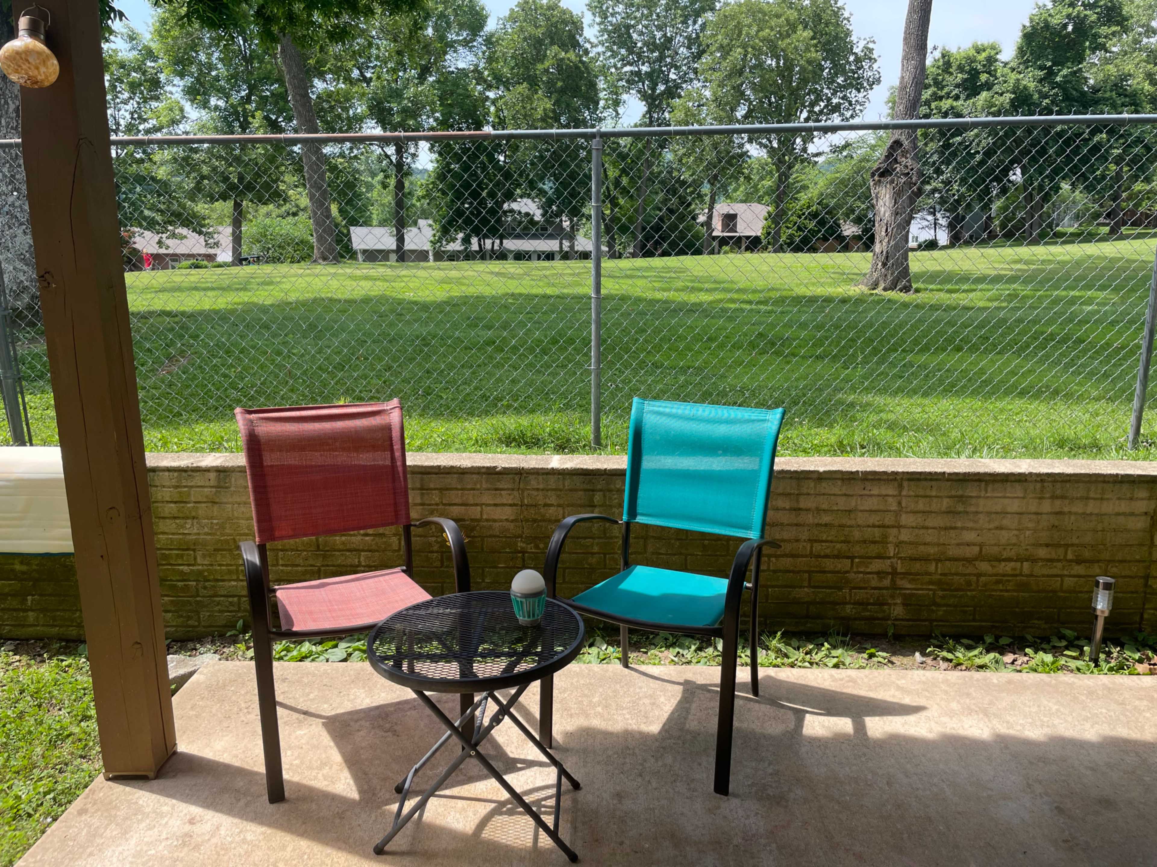 Two chairs, one red and one teal, are situated beside a small table on a patio overlooking a green yard enclosed by a chain-link fence.