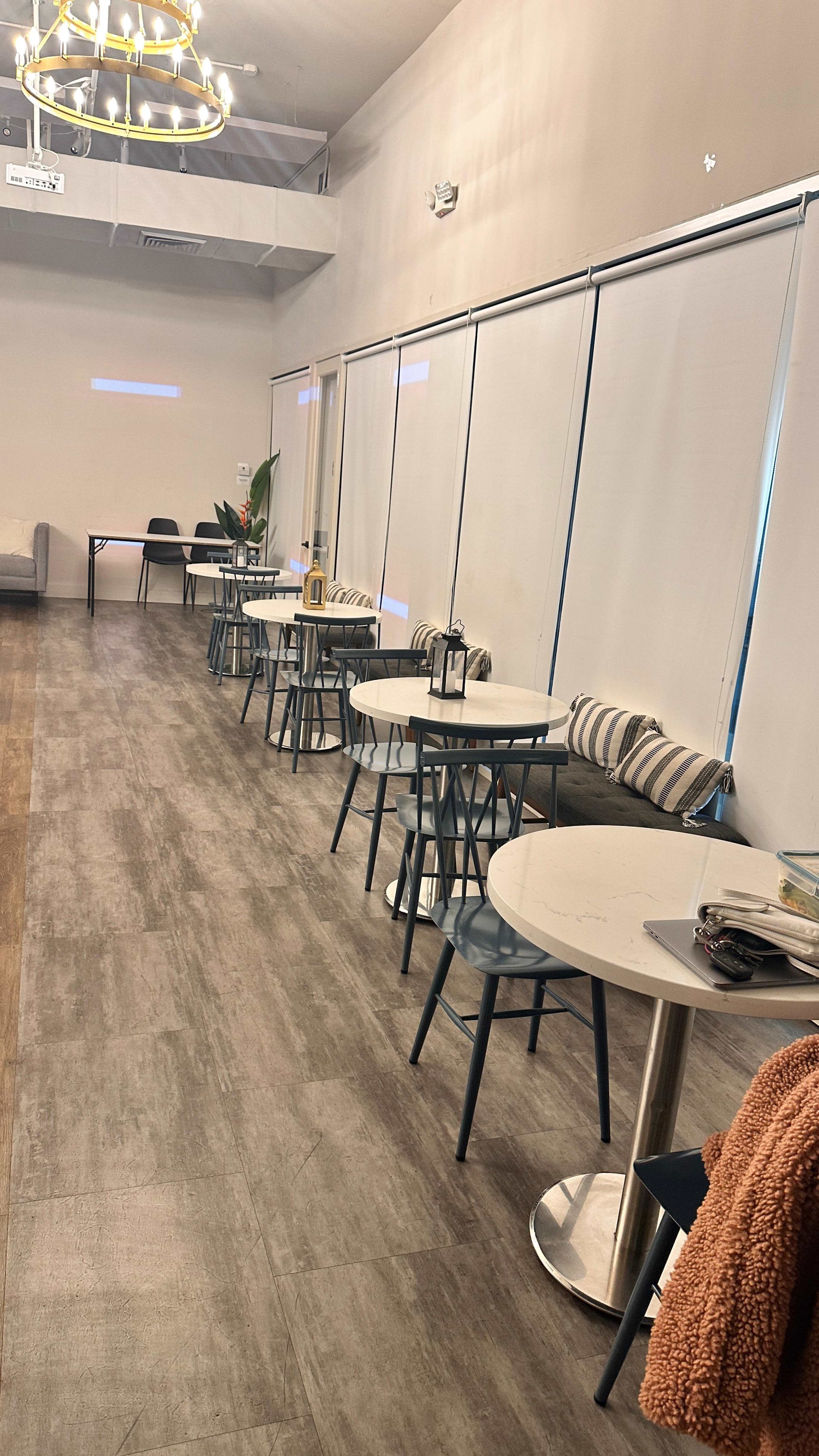 The image shows a spacious interior with several small tables and chairs aligned along a wall, featuring large windows covered by shades and a modern light fixture hanging from the ceiling.