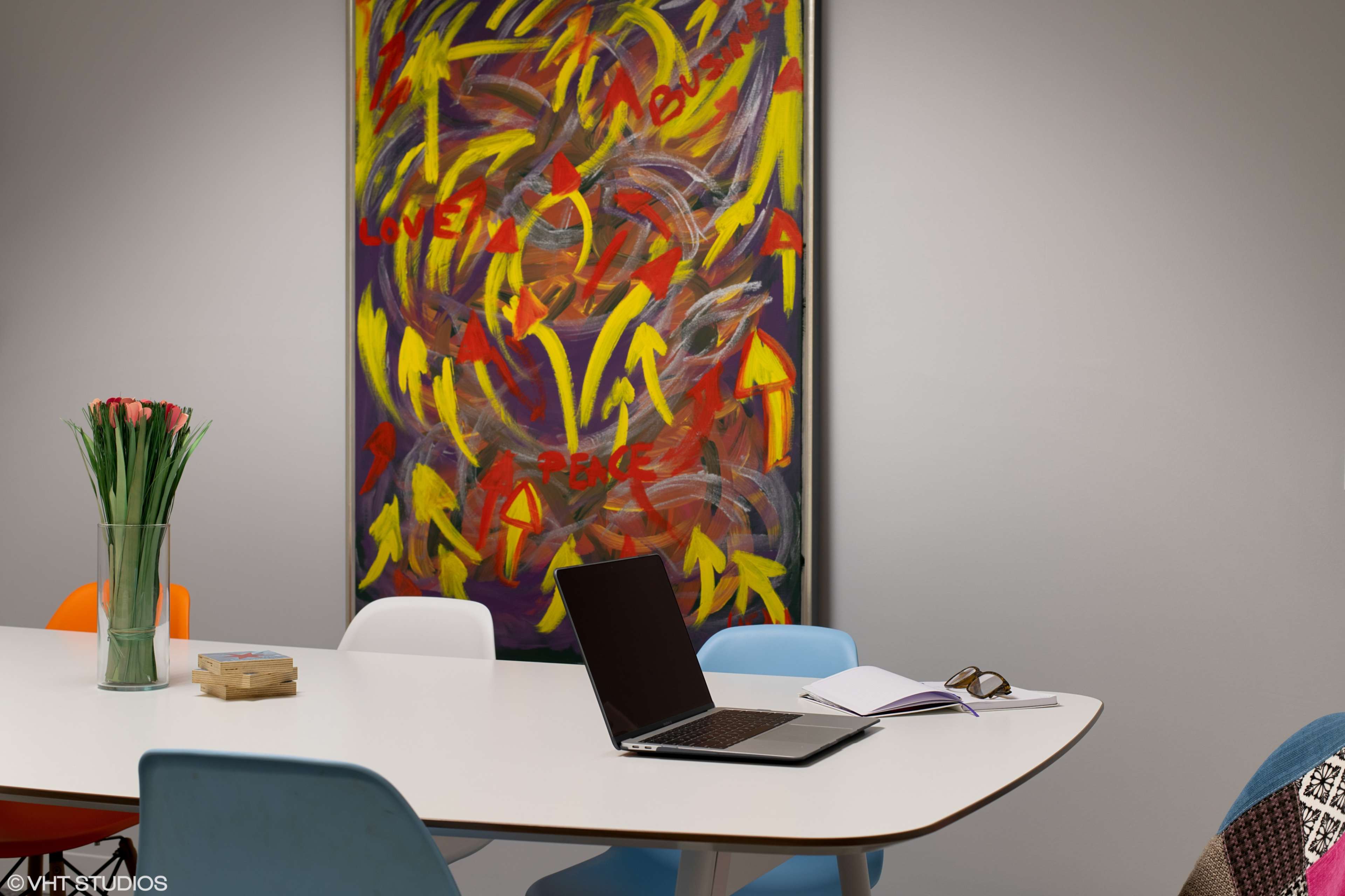 A modern conference room features a round table with colorful chairs, a laptop, and an open notebook, alongside a vibrant abstract painting on the wall.