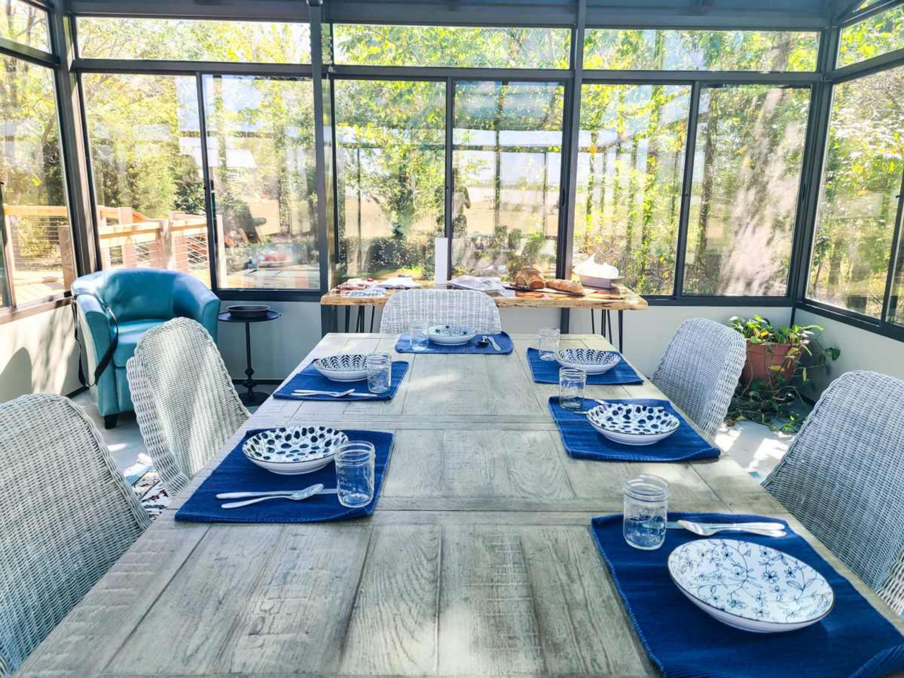 A dining table is set for a meal in a sunroom with large windows, surrounded by chairs and decor.
