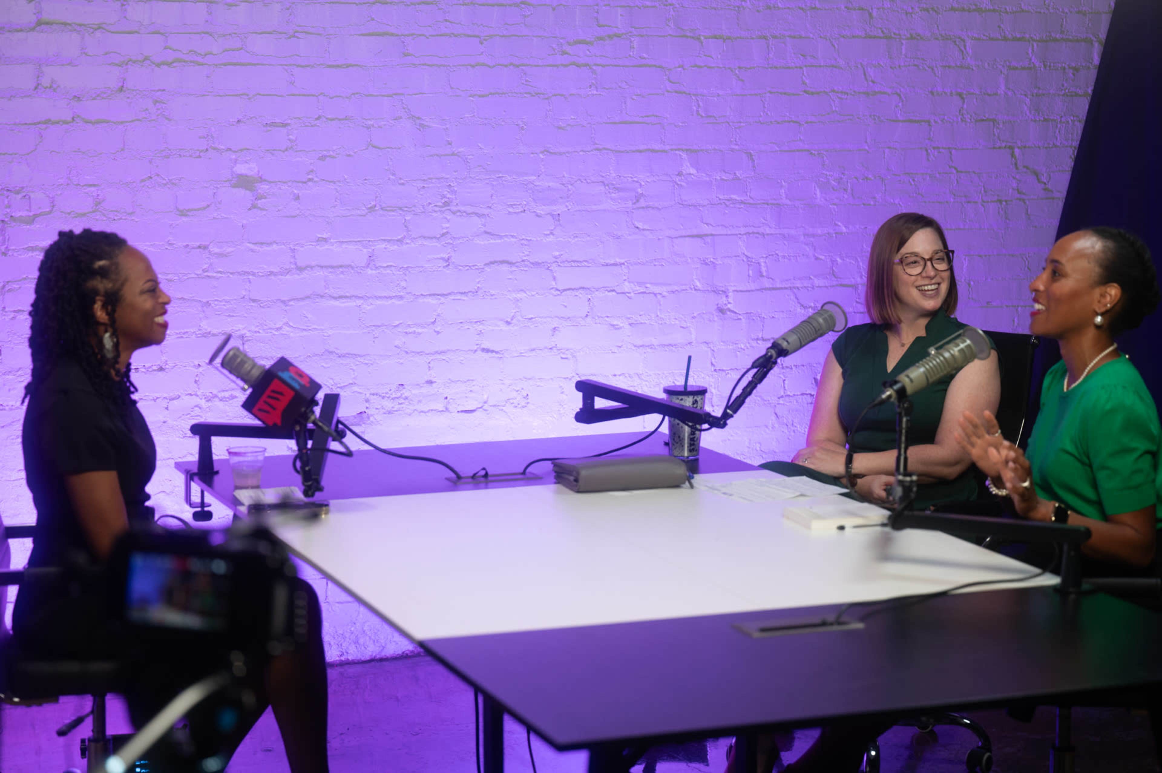 Three women sit at a table equipped with microphones in a well-lit studio with a purple backdrop, engaged in conversation.
