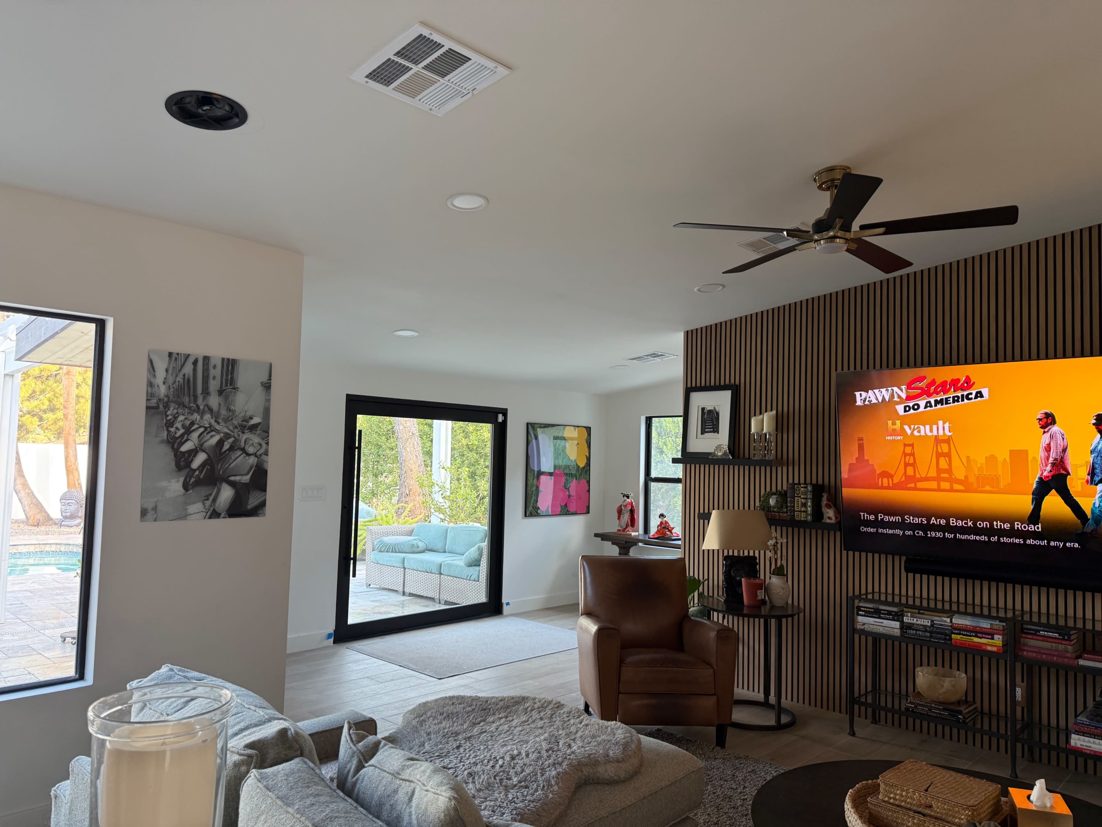 A modern living room features a television displaying a show, a brown armchair, and a view of an outdoor space through large glass doors.