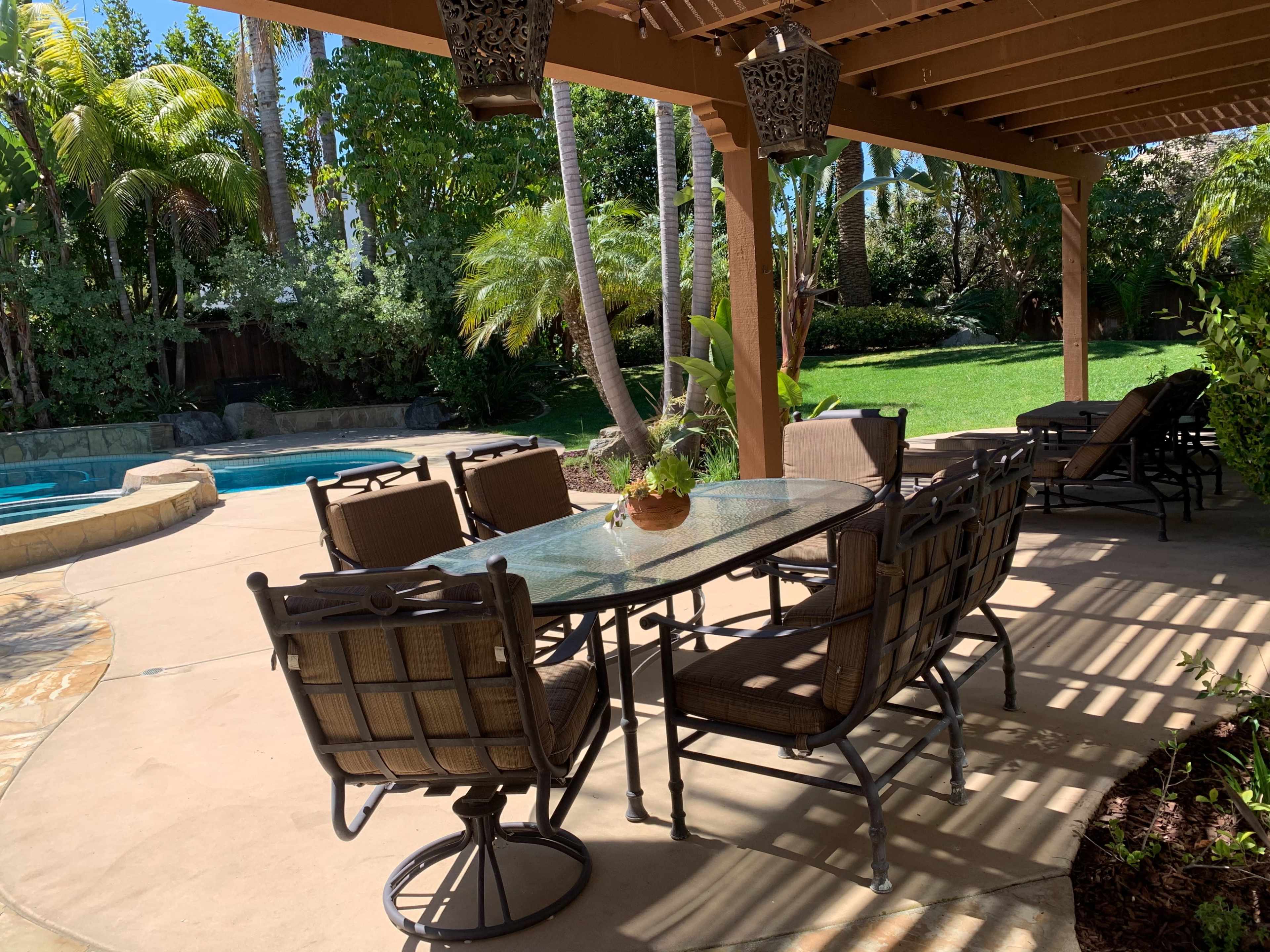 A backyard patio features a glass dining table surrounded by chairs, with a swimming pool and lush greenery in the background.