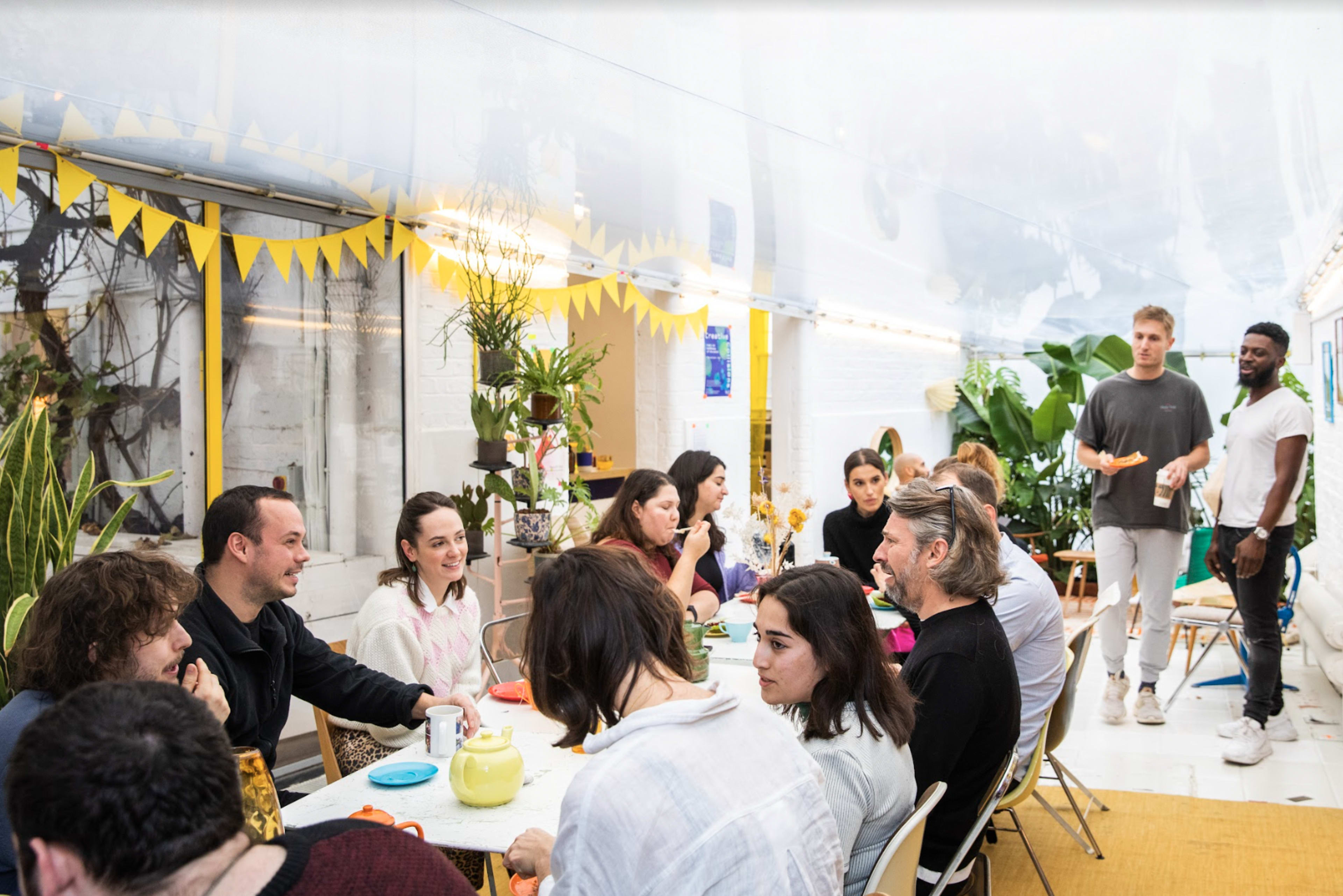 A group of people gathers around tables in a bright, decorated room filled with plants and vibrant decorations.