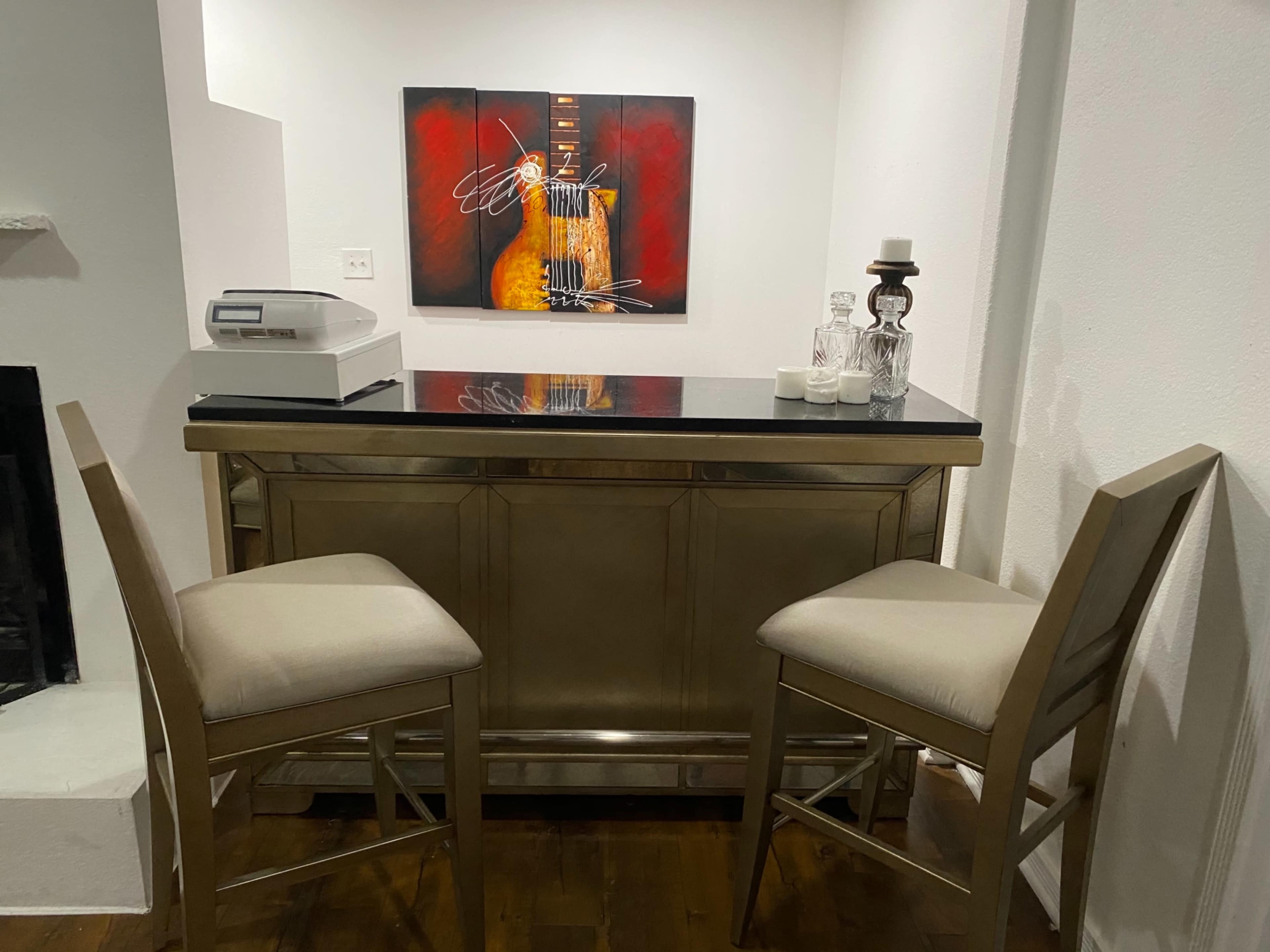 The image shows a small bar area with two beige chairs, a black countertop, a printer, and an abstract painting of a guitar on the wall.