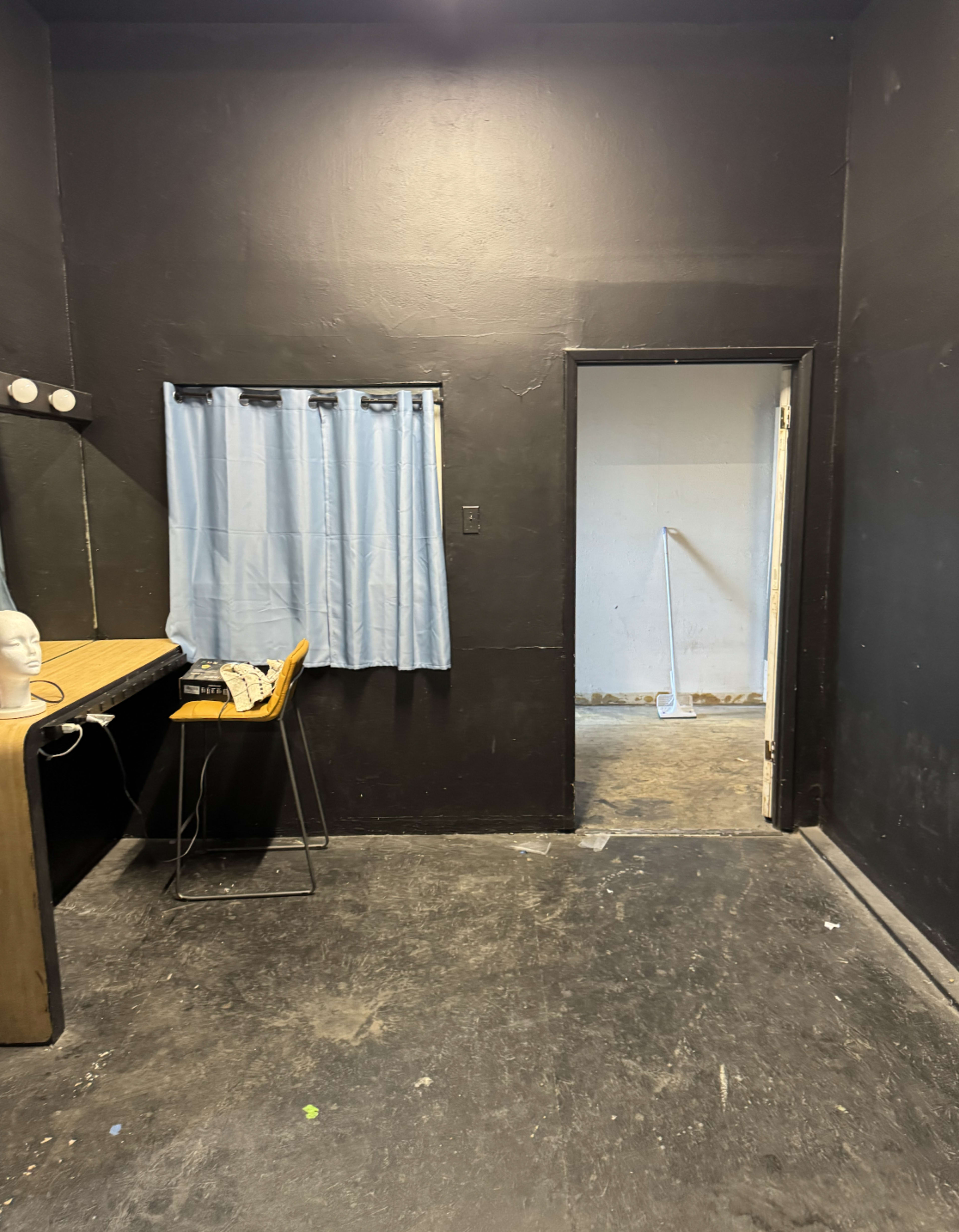 The image shows a dimly lit room with black walls, a small window covered by a light blue curtain, a table with a mirror, and an open door leading to another space.