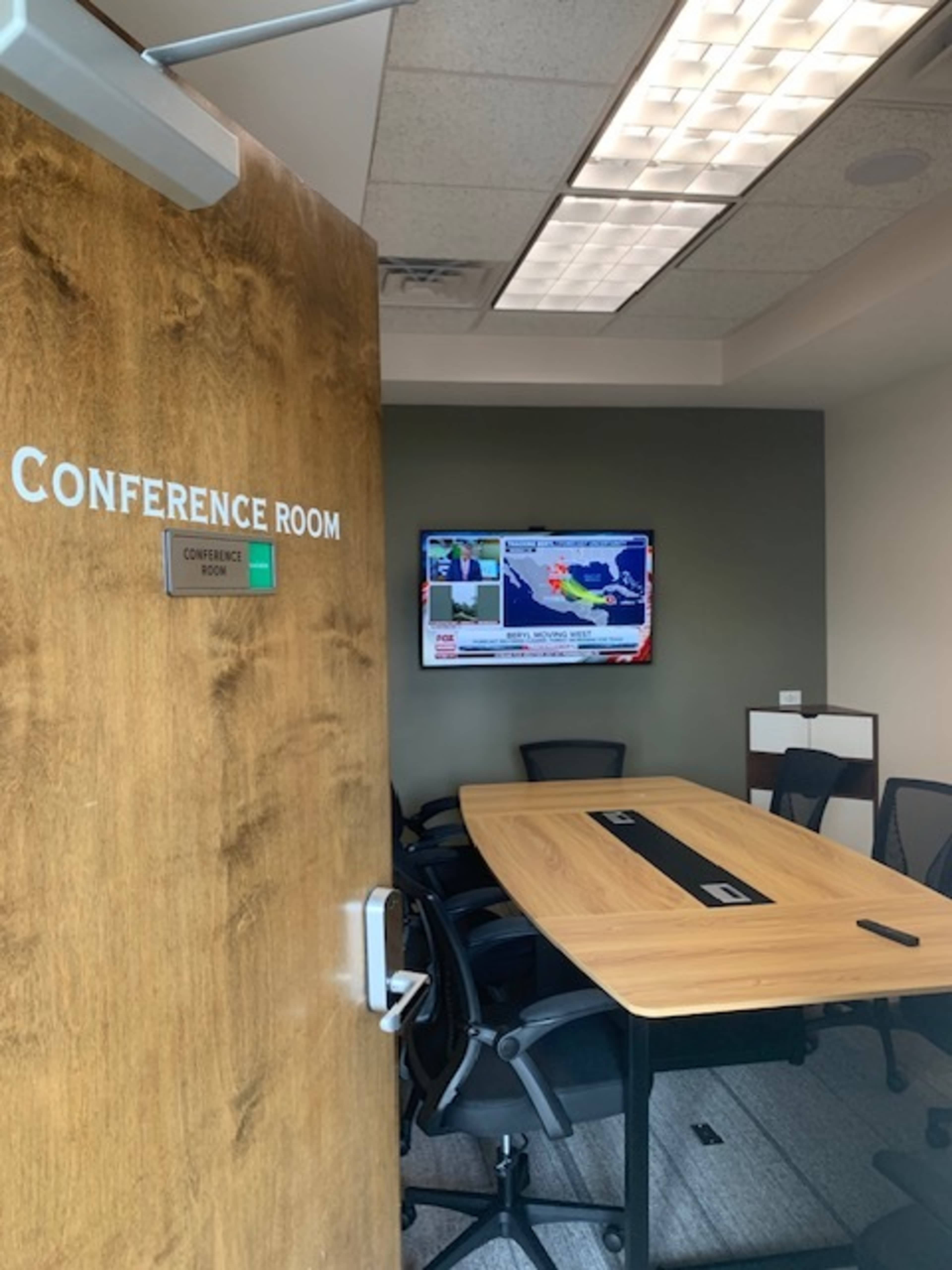 A conference room is visible through an open door, featuring a wooden table and a television displaying a weather report.
