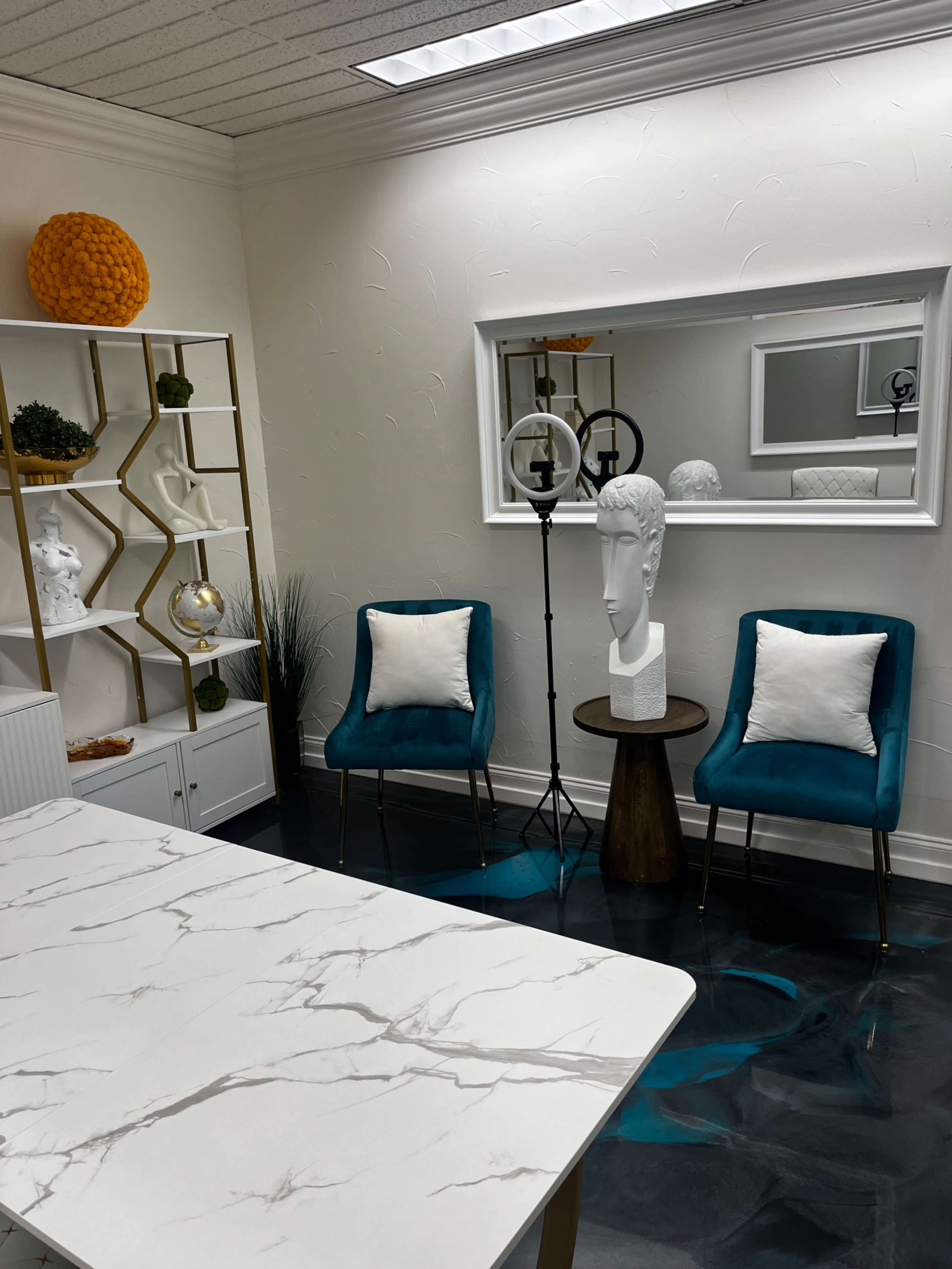 The image shows a well-decorated, modern interior space with two teal chairs, a side table, a decorative mirror, a bookshelf, and various ornaments.