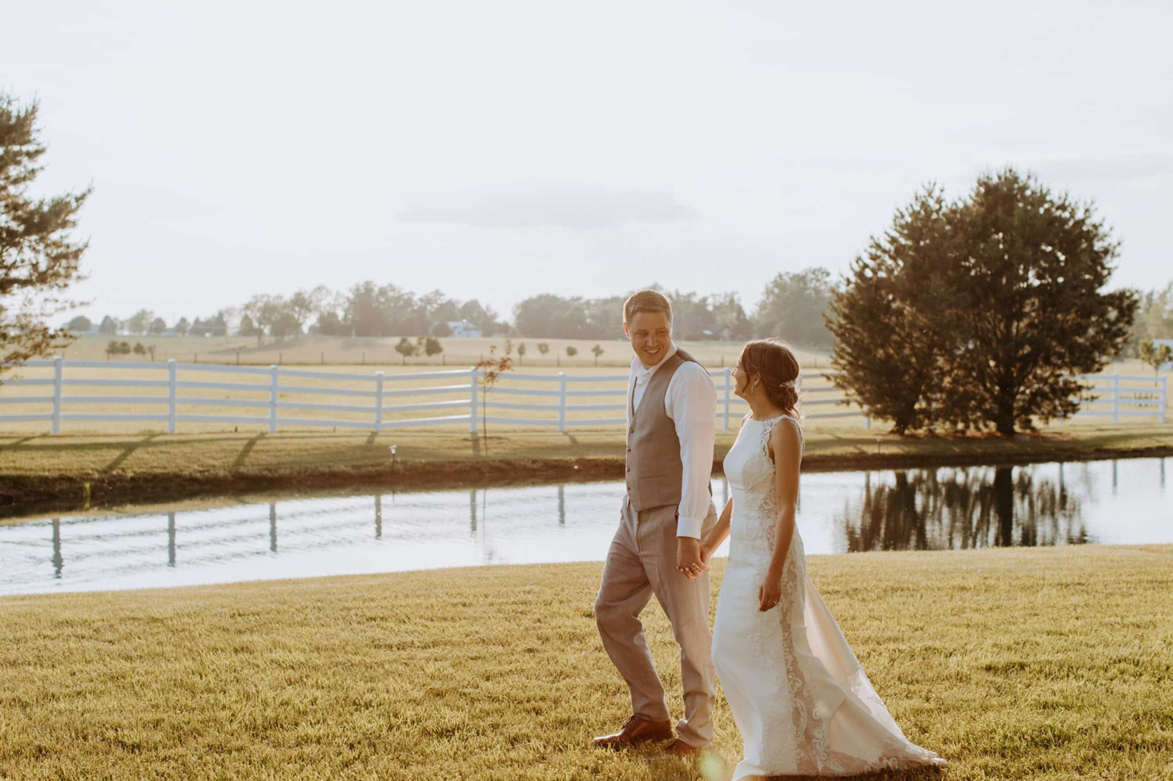 A bride and groom walk hand in hand along a grassy area near a pond, with a white fence and trees in the background.