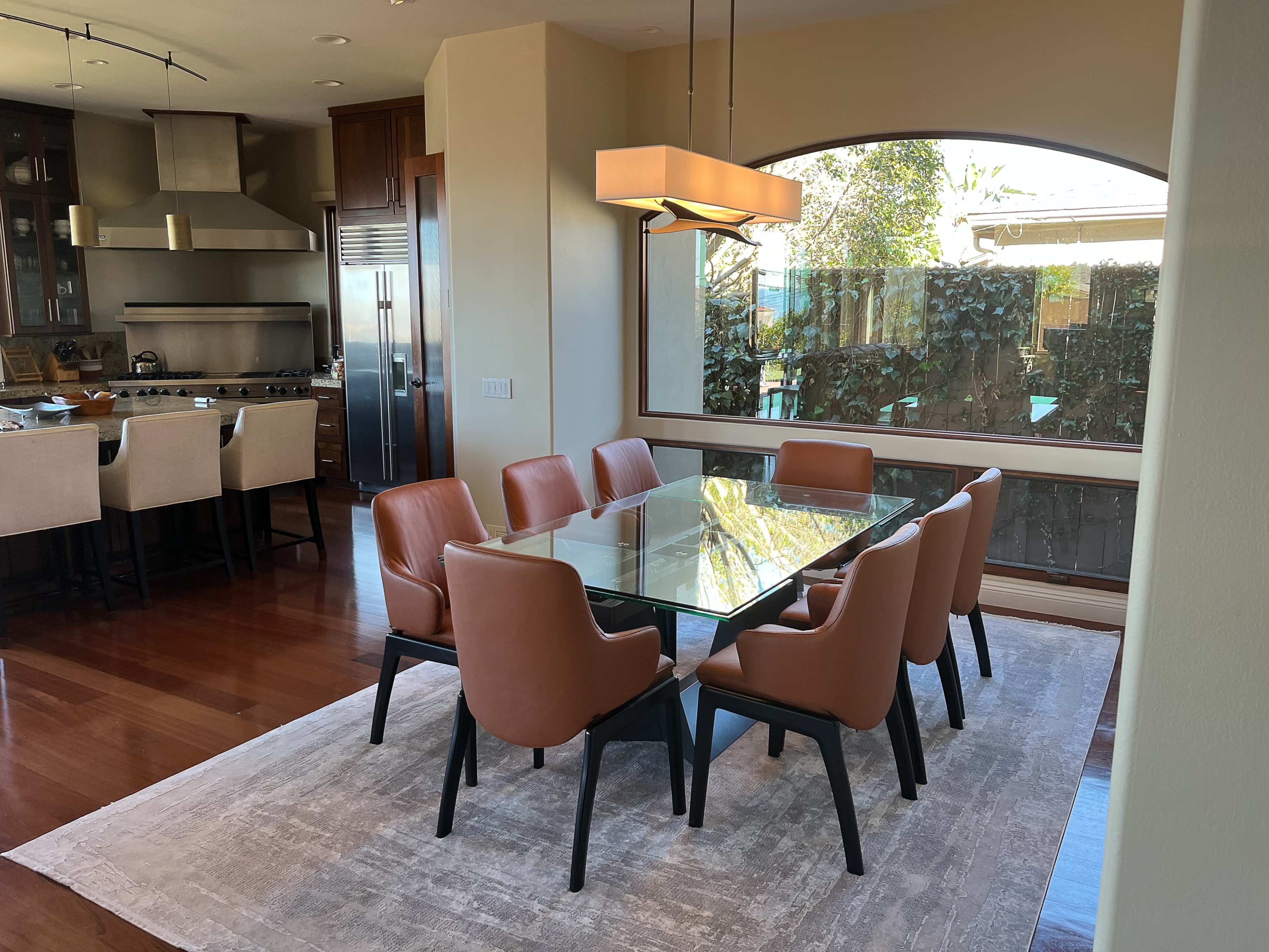 A glass dining table surrounded by brown chairs is positioned on a rug in a well-lit dining area that opens to a garden view.