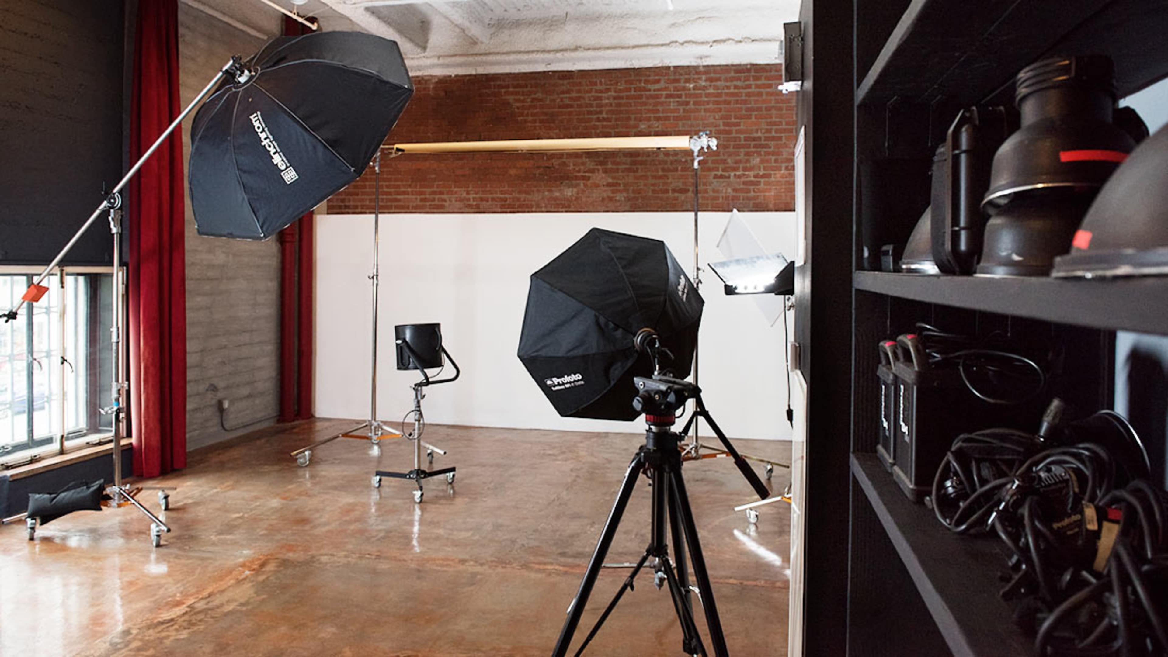 The image shows a photography studio equipped with various lighting setups, a backdrop support, and camera equipment on a shelf.