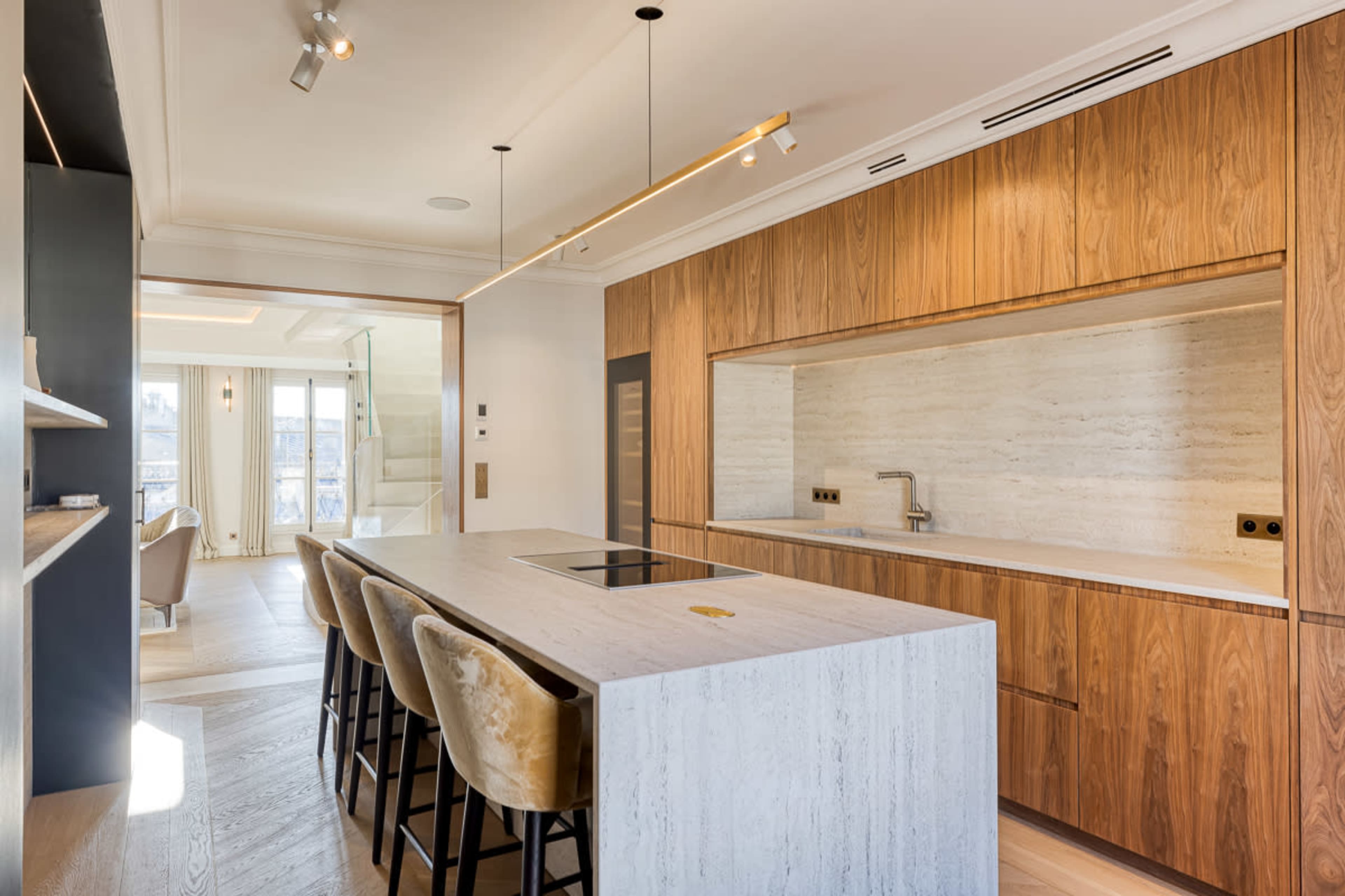 The image shows a modern kitchen with a large island, featuring wooden cabinetry, a stone countertop, and stylish bar stools.