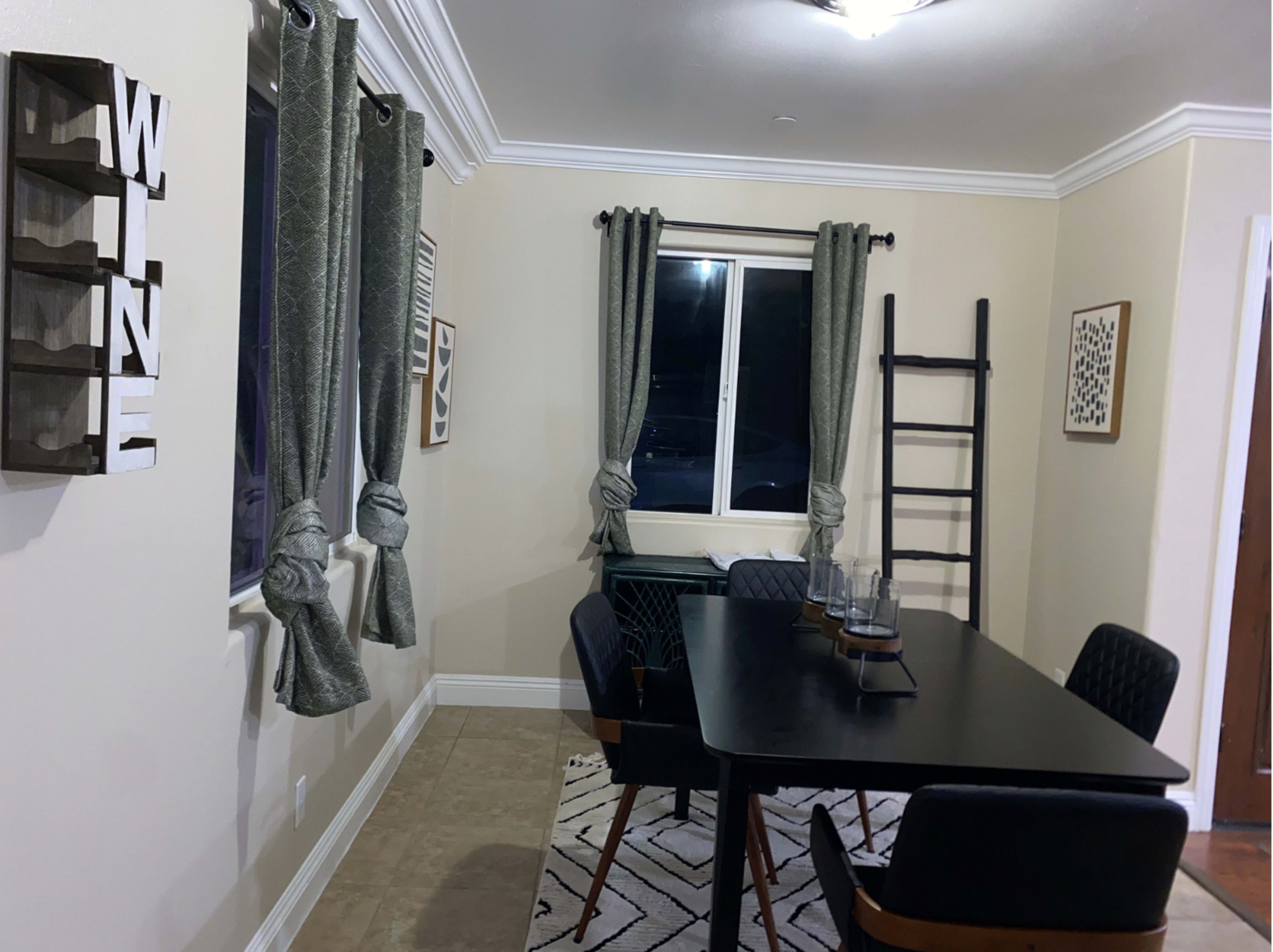 A dining area features a black table surrounded by chairs, with curtains framing a window and a decorative ladder against the wall.