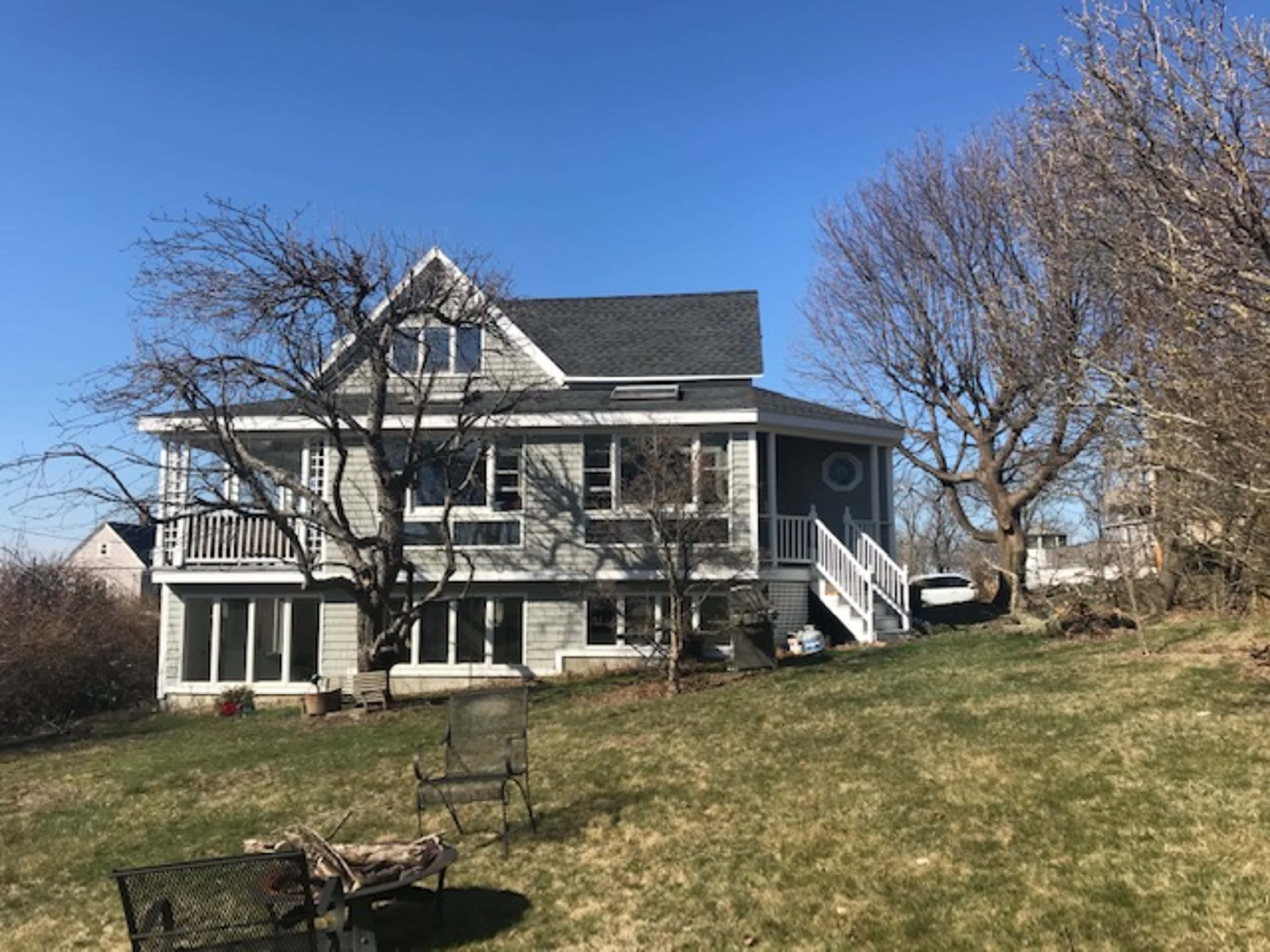 A two-story house with a gray exterior and a large yard featuring bare trees and a few outdoor chairs.