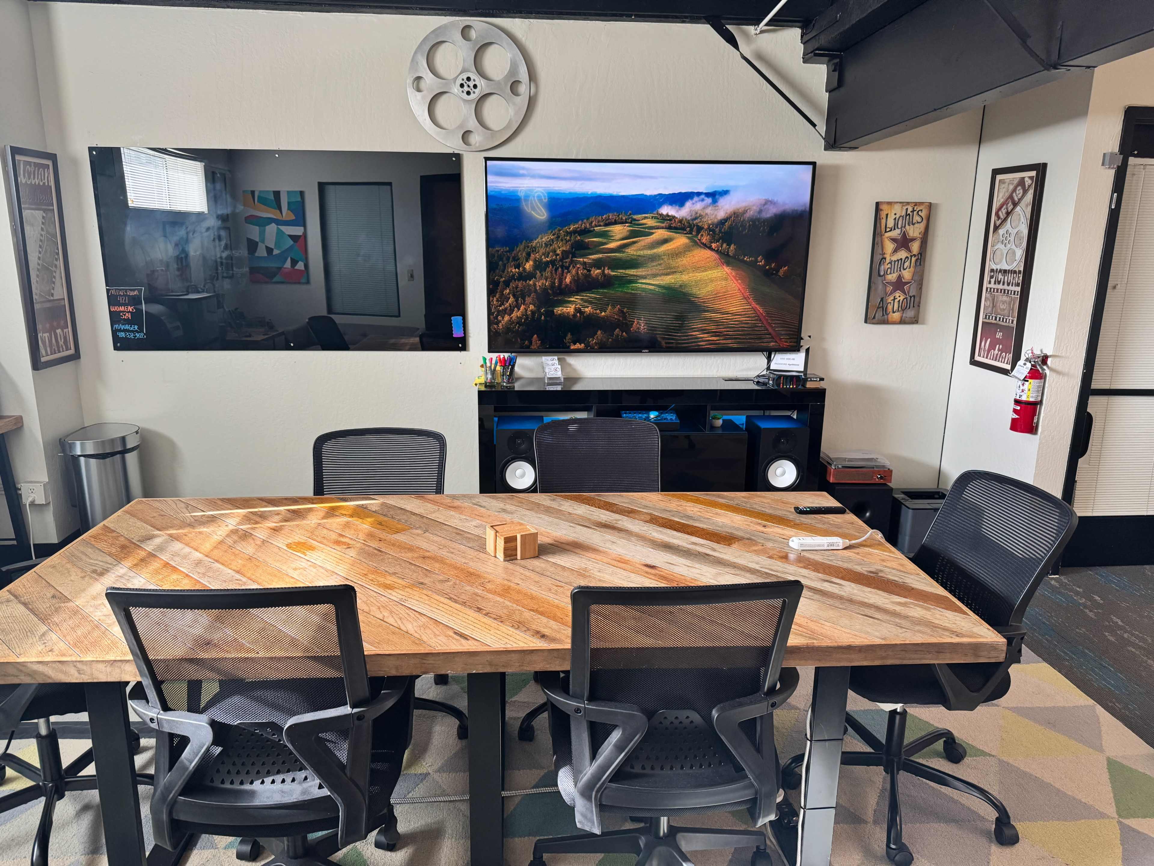 The image shows a modern conference room with a large wooden table, several black office chairs, and two large screens mounted on the wall displaying a landscape and tools.