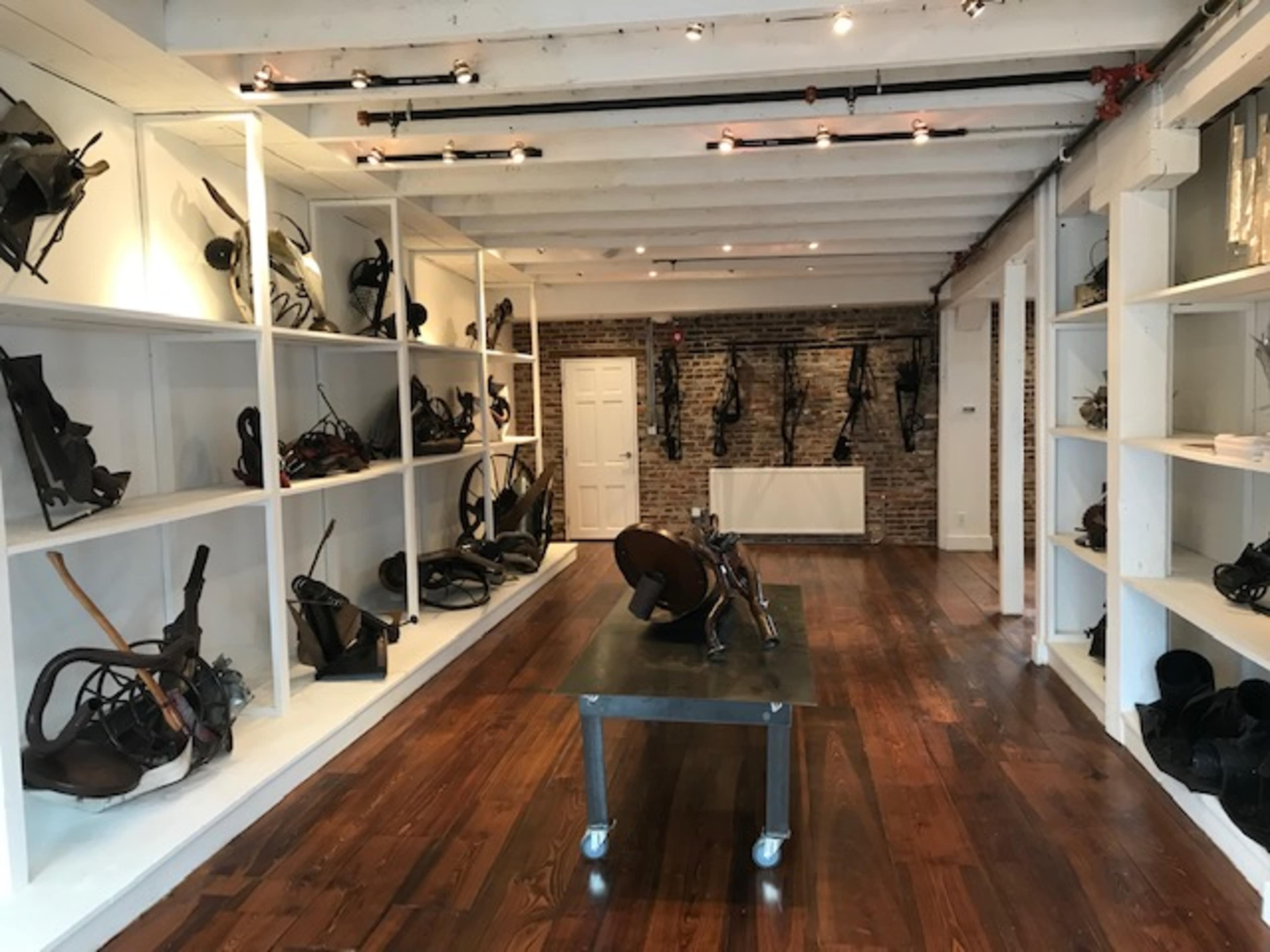The image shows an art gallery with shelves filled with various metal sculptures and tools, set against a brick wall and wooden flooring.