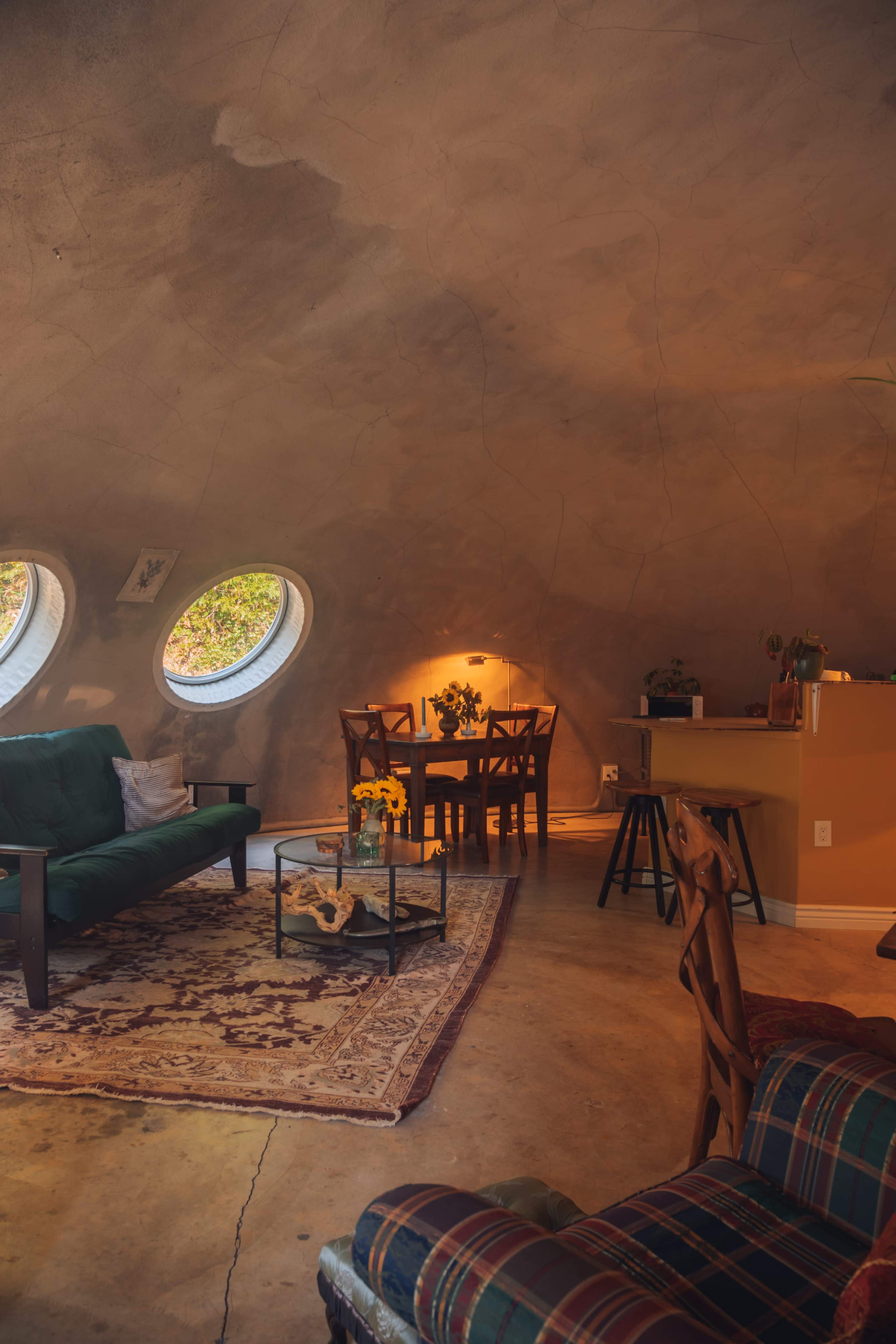 The image shows a cozy living space with a round, textured ceiling, a green sofa, a dining area with two chairs and a small table, and large circular windows letting in natural light.