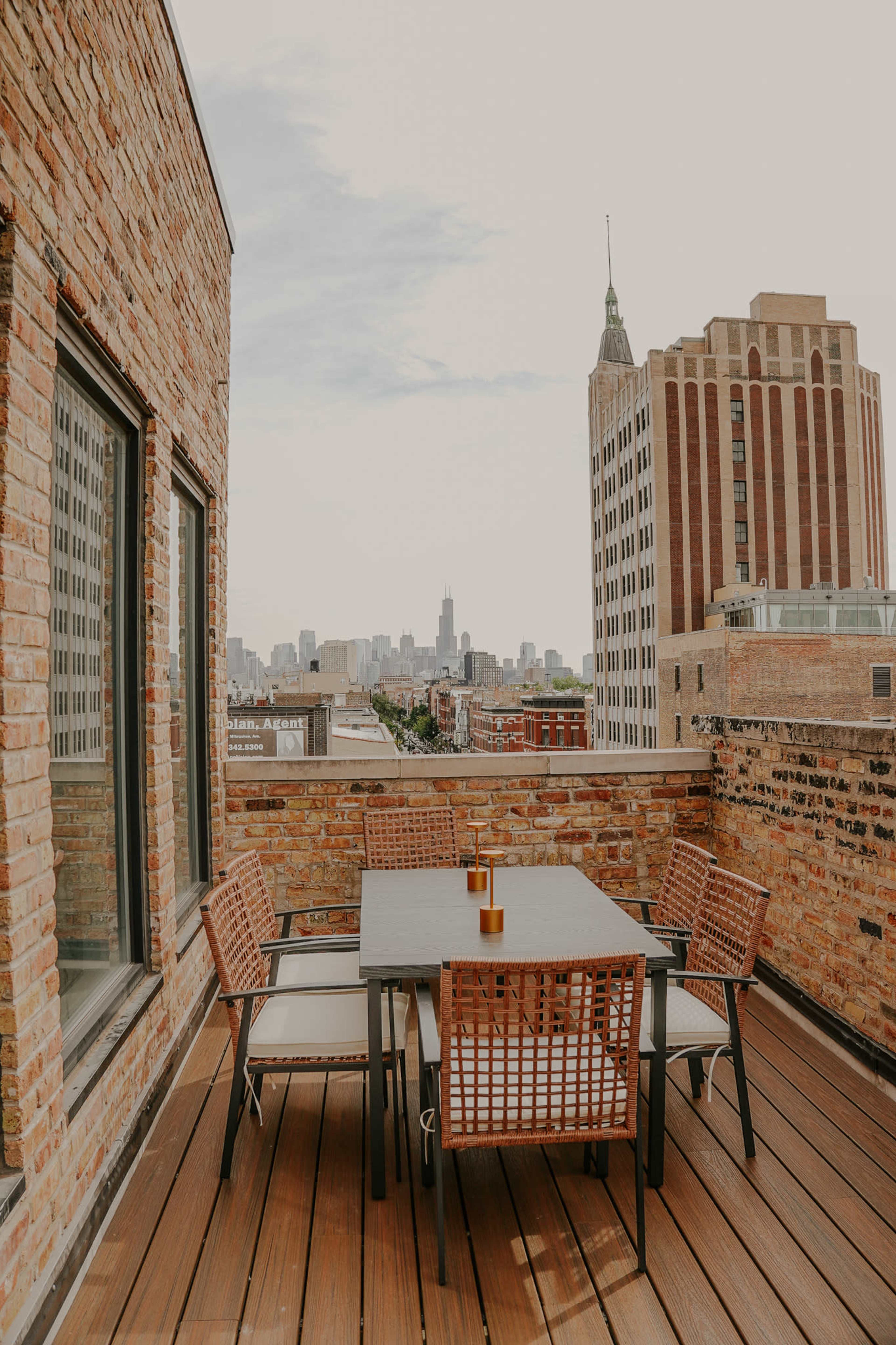 A rooftop patio features a dining table with chairs and offers a view of a city skyline, including notable buildings.