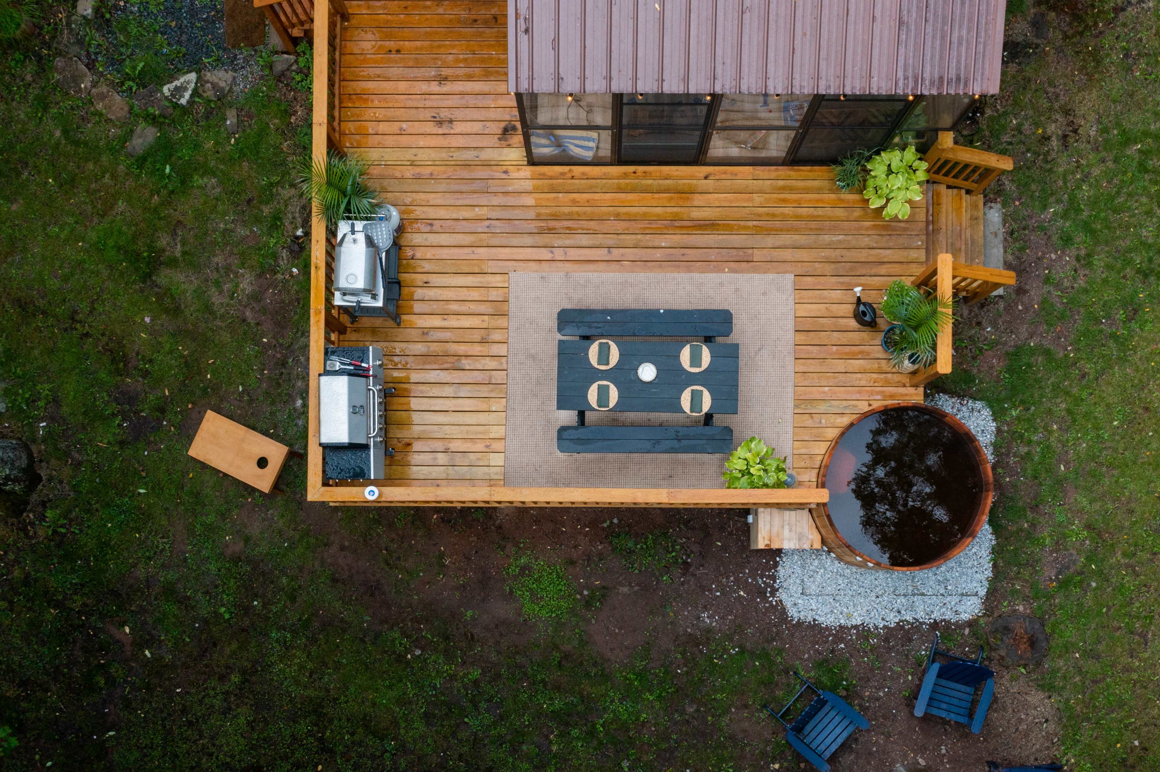 An aerial view shows a wooden deck with a rectangular dining table, a grill, potted plants, and a small circular water feature.