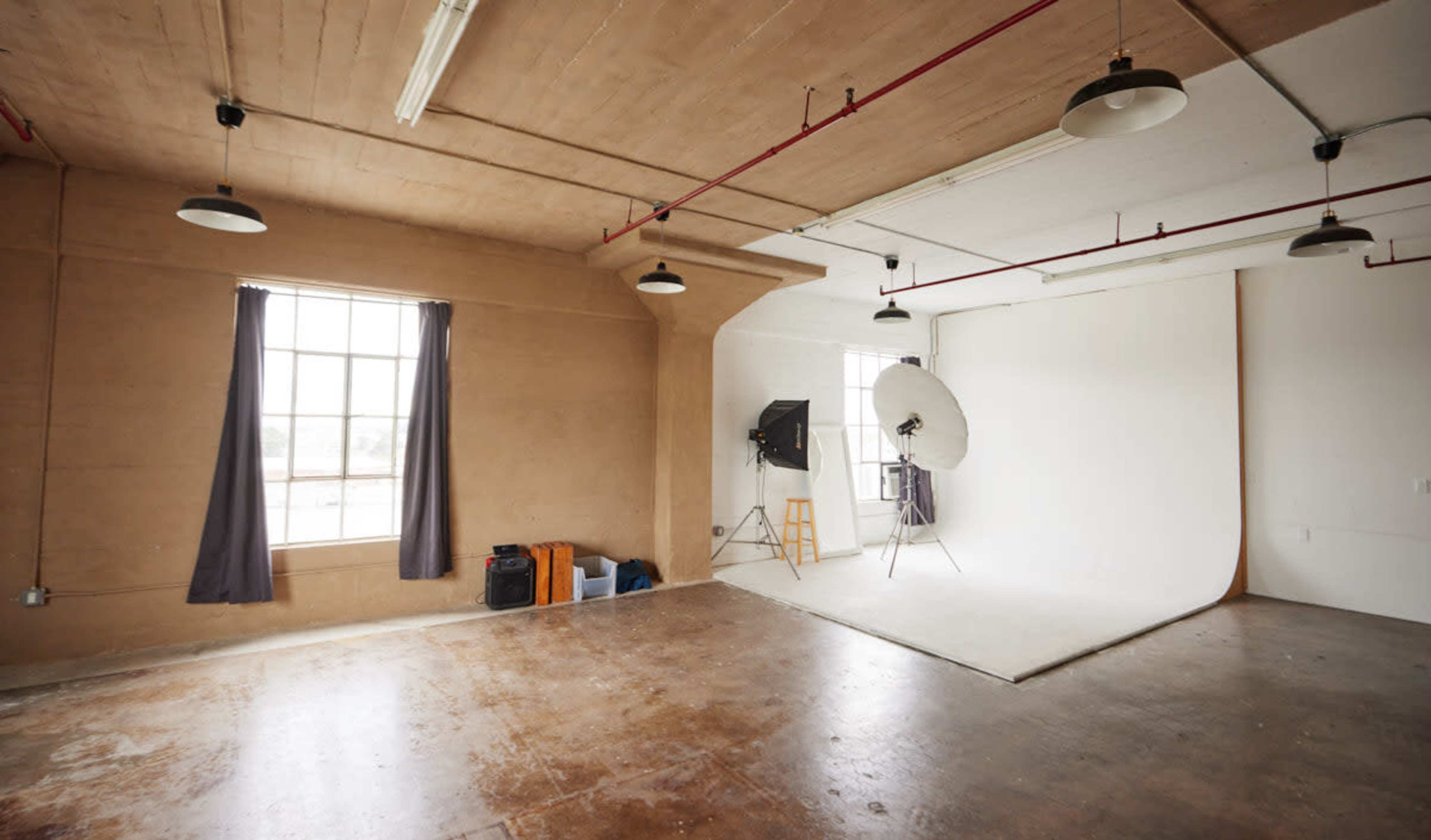 An empty photography studio with a large light umbrella and backdrop, illuminated by overhead lights and natural light from a window.