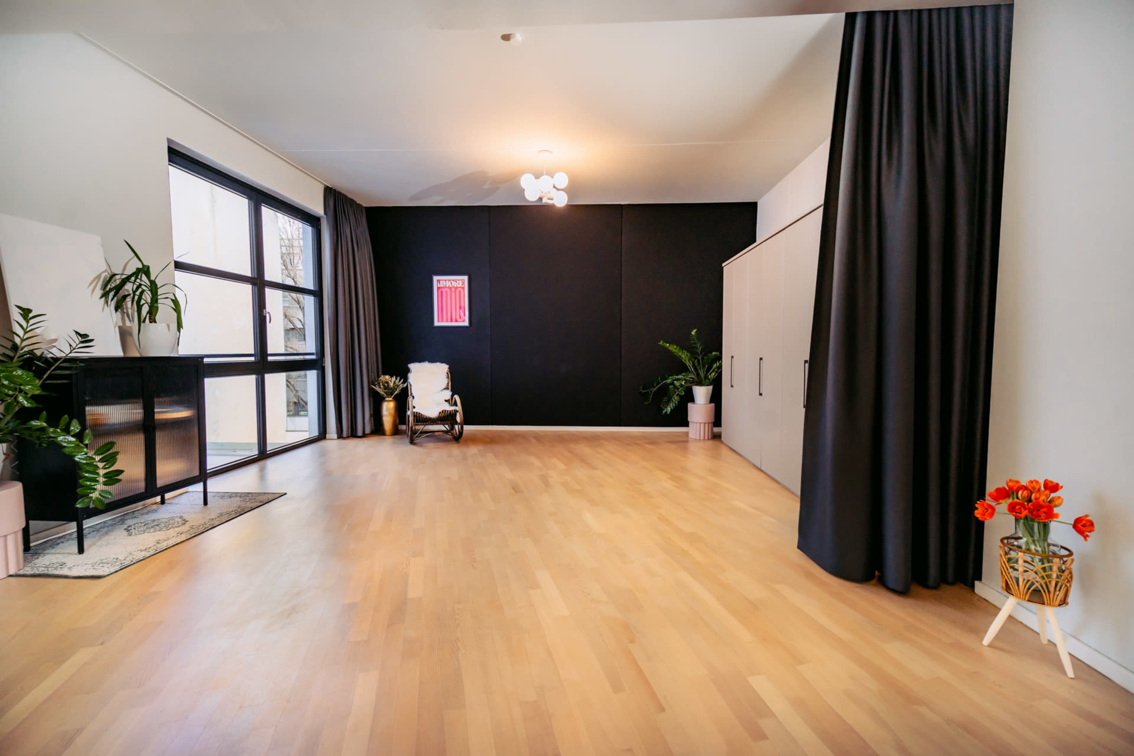 A spacious room with wooden flooring, large windows, black curtains, a modern light fixture, and a few decorative plants.